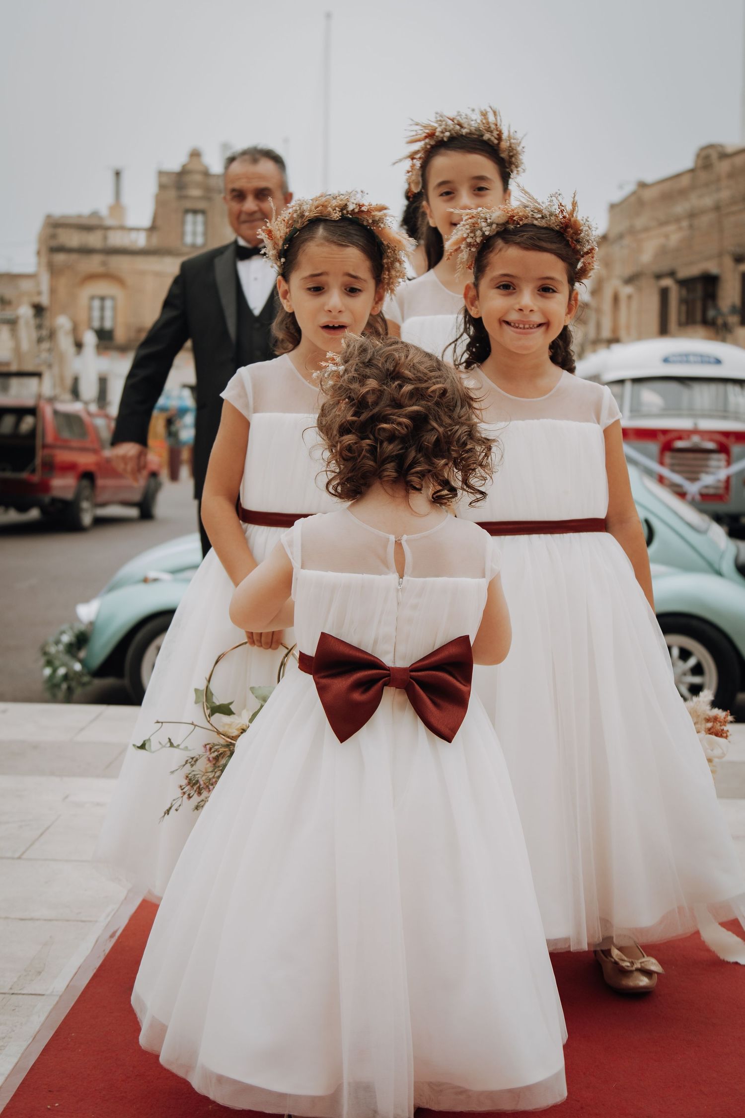 Flower girls in white dresses with burgundy sashes gather on red carpet outside historic building.
