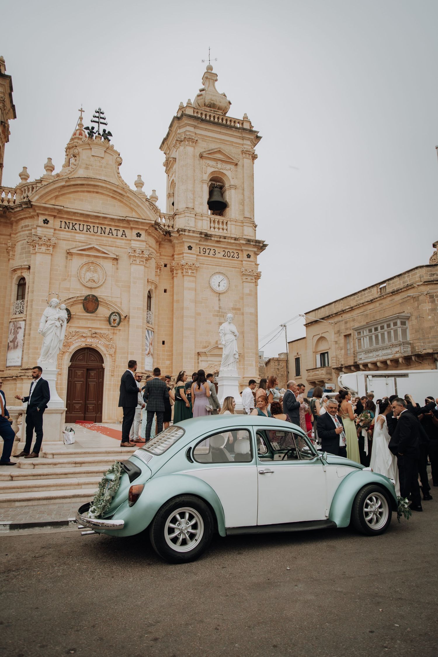 Baroque church facade with classic VW Beetle and wedding guests gathered on the steps.