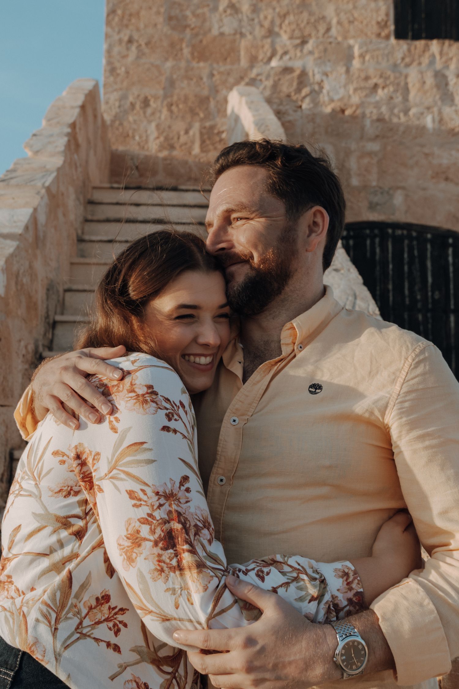 Intimate couple shares tender moments while wearing cream and floral outfits against stone wall backdrop.