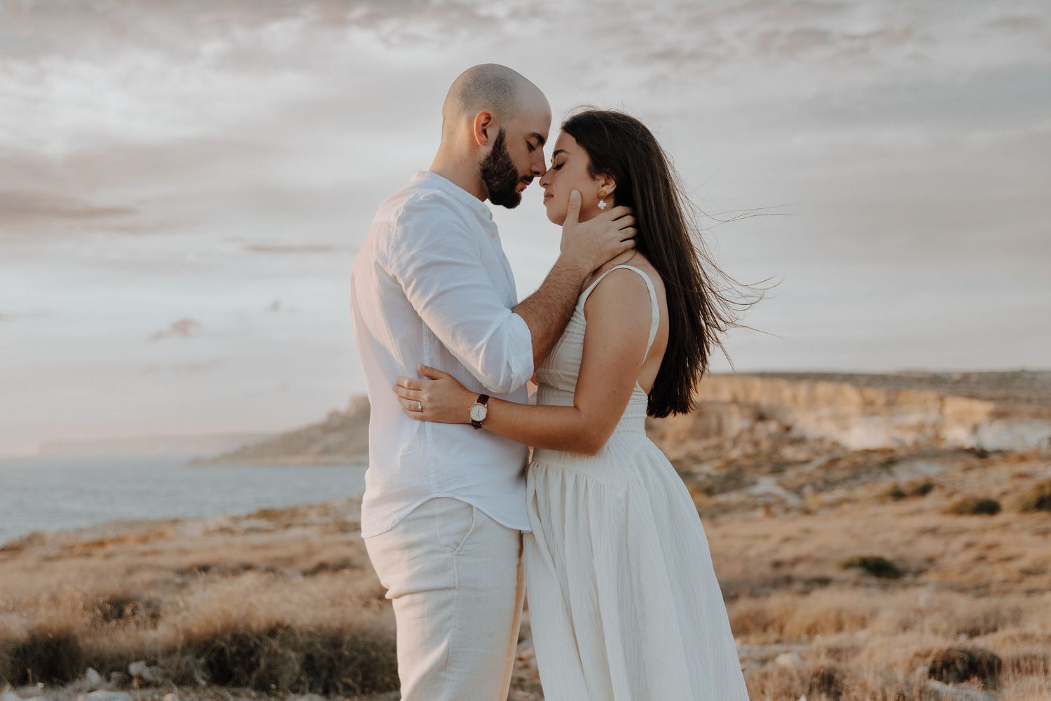 A romantic couple in white outfits embrace on a coastal cliff during sunset with ocean views in the background.
