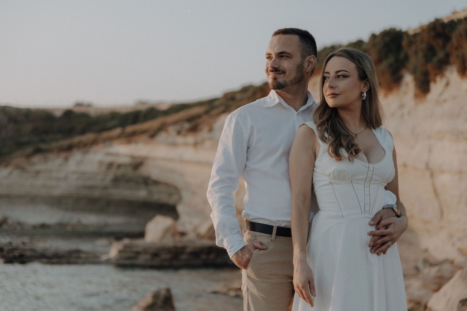 Couple in white attire embracing near rocky coastal cliffs at golden hour sunset.