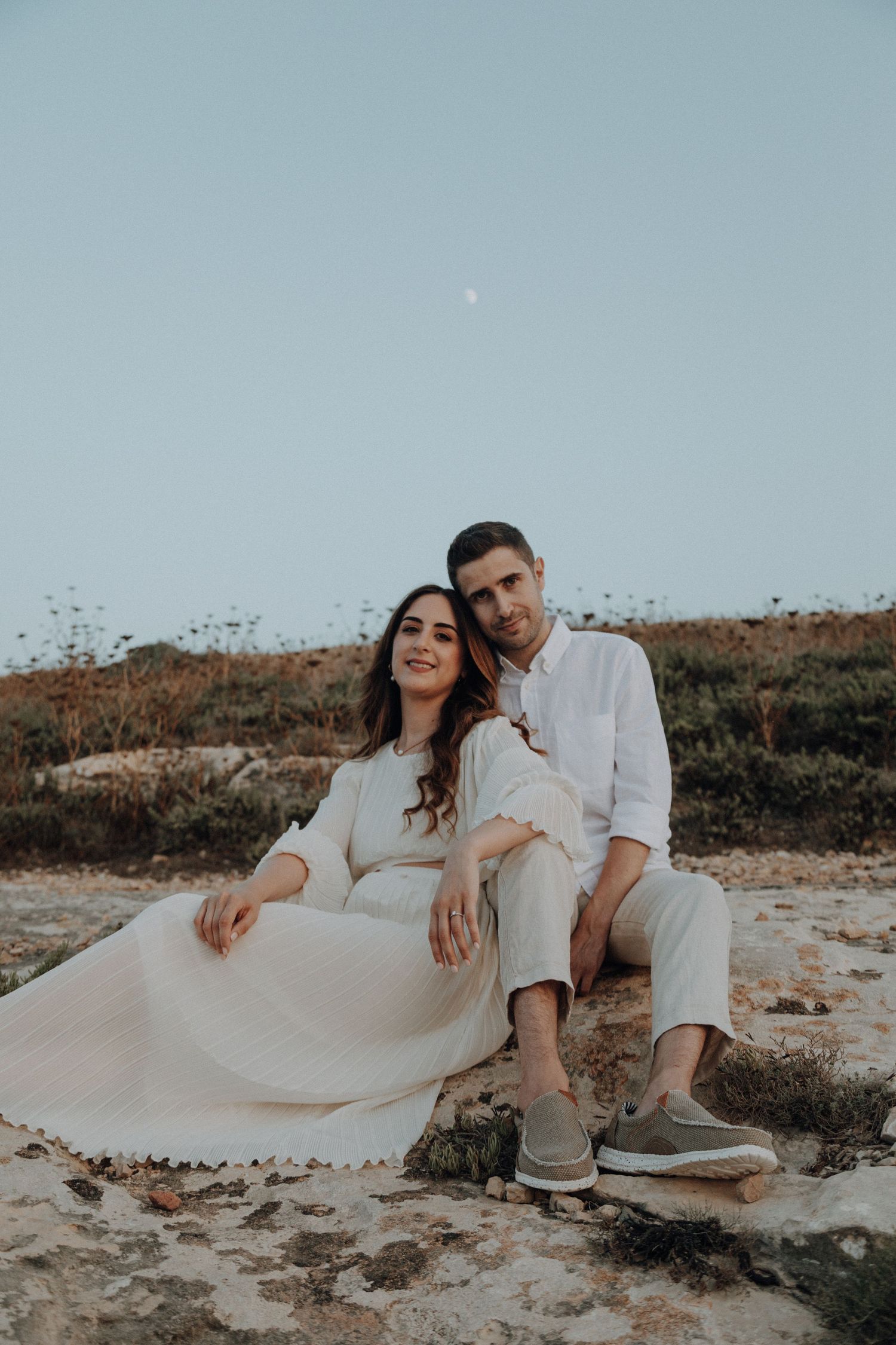 Couple in matching white outfits sitting on coastal rocks during golden hour.