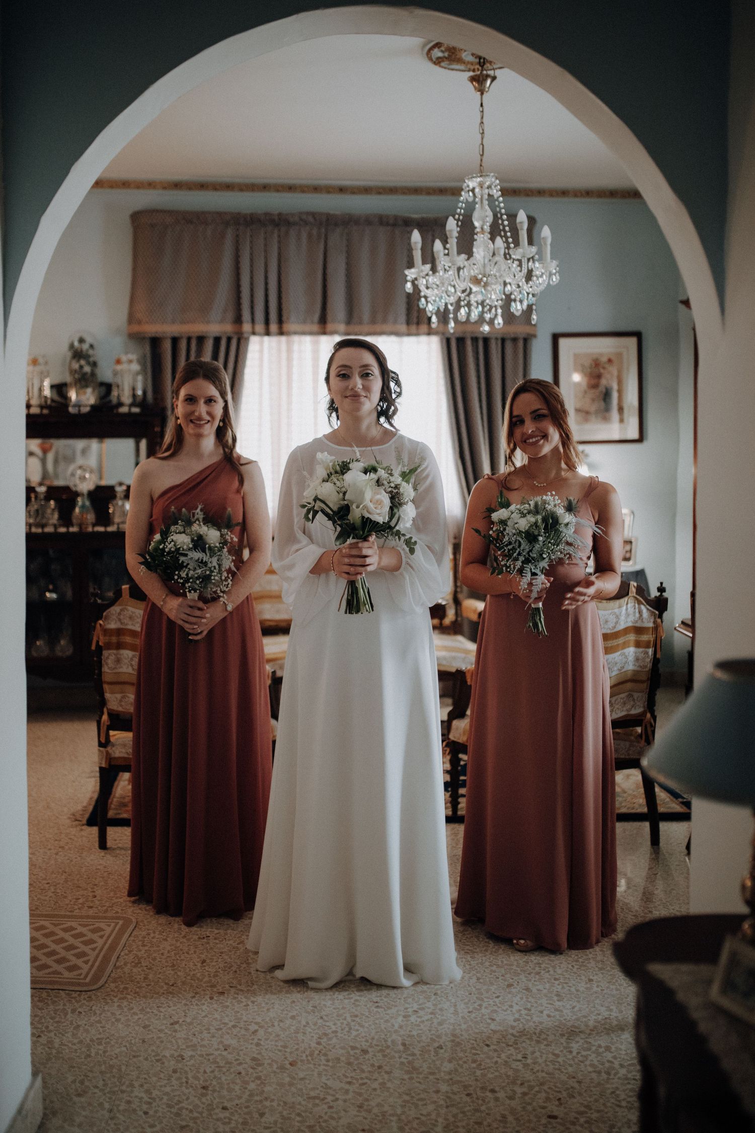 Bridal party poses in white and burgundy dresses under an elegant archway with chandelier.