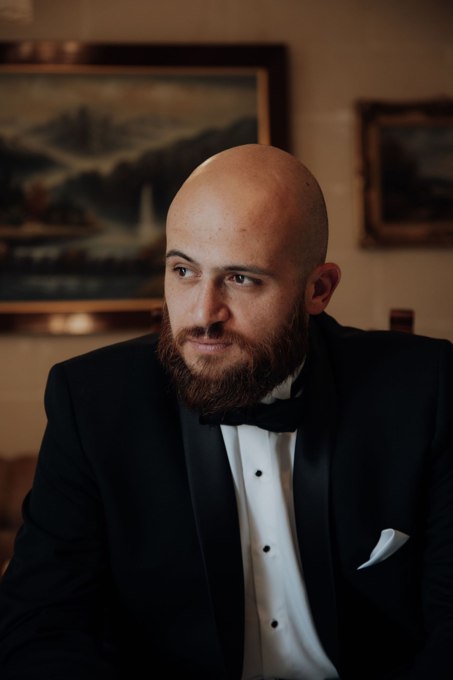Man in tuxedo and bow tie sitting thoughtfully in formal setting.