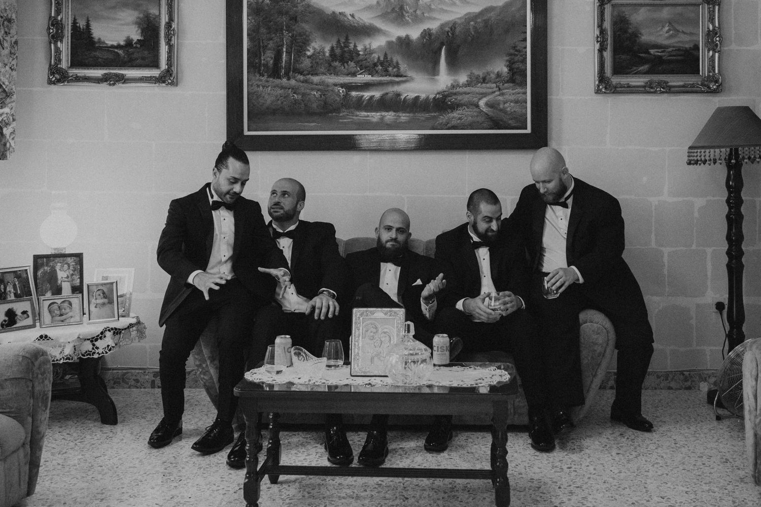 Black and white photo of groomsmen relaxing on vintage furniture before wedding ceremony.