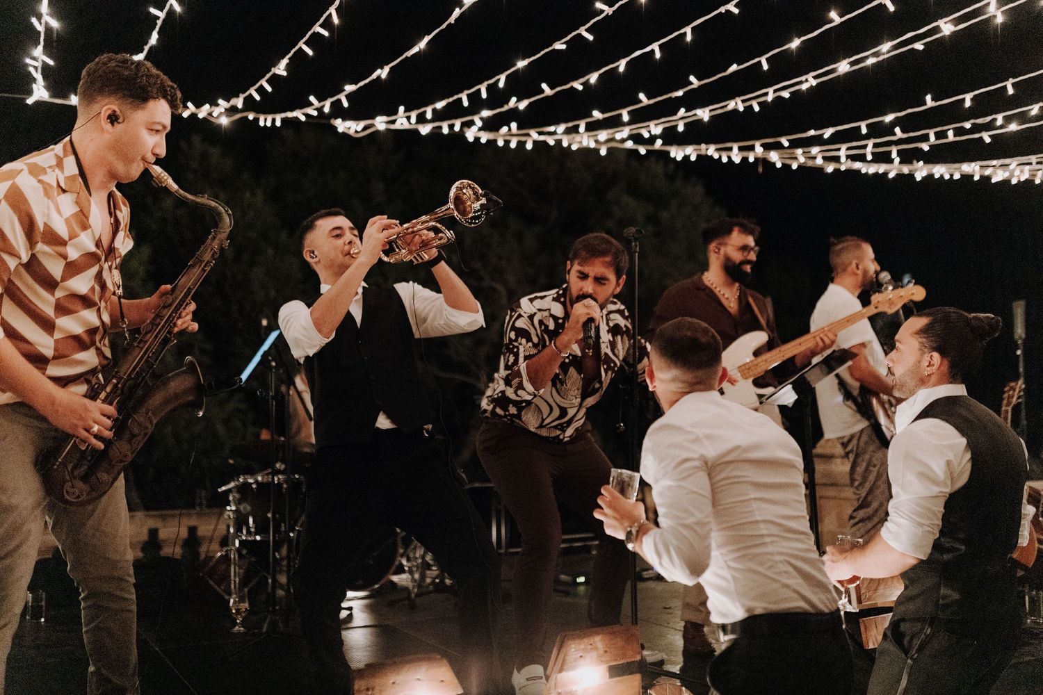 Musicians playing instruments at a wedding reception under string lights create a lively atmosphere.