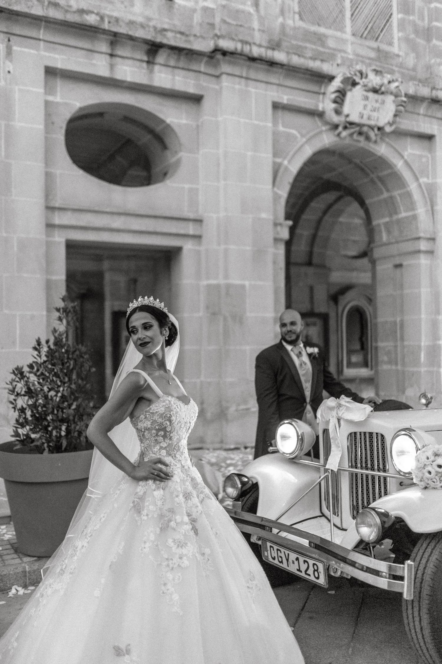 Black and white portrait of bride posing elegantly by vintage car with groom in background.
