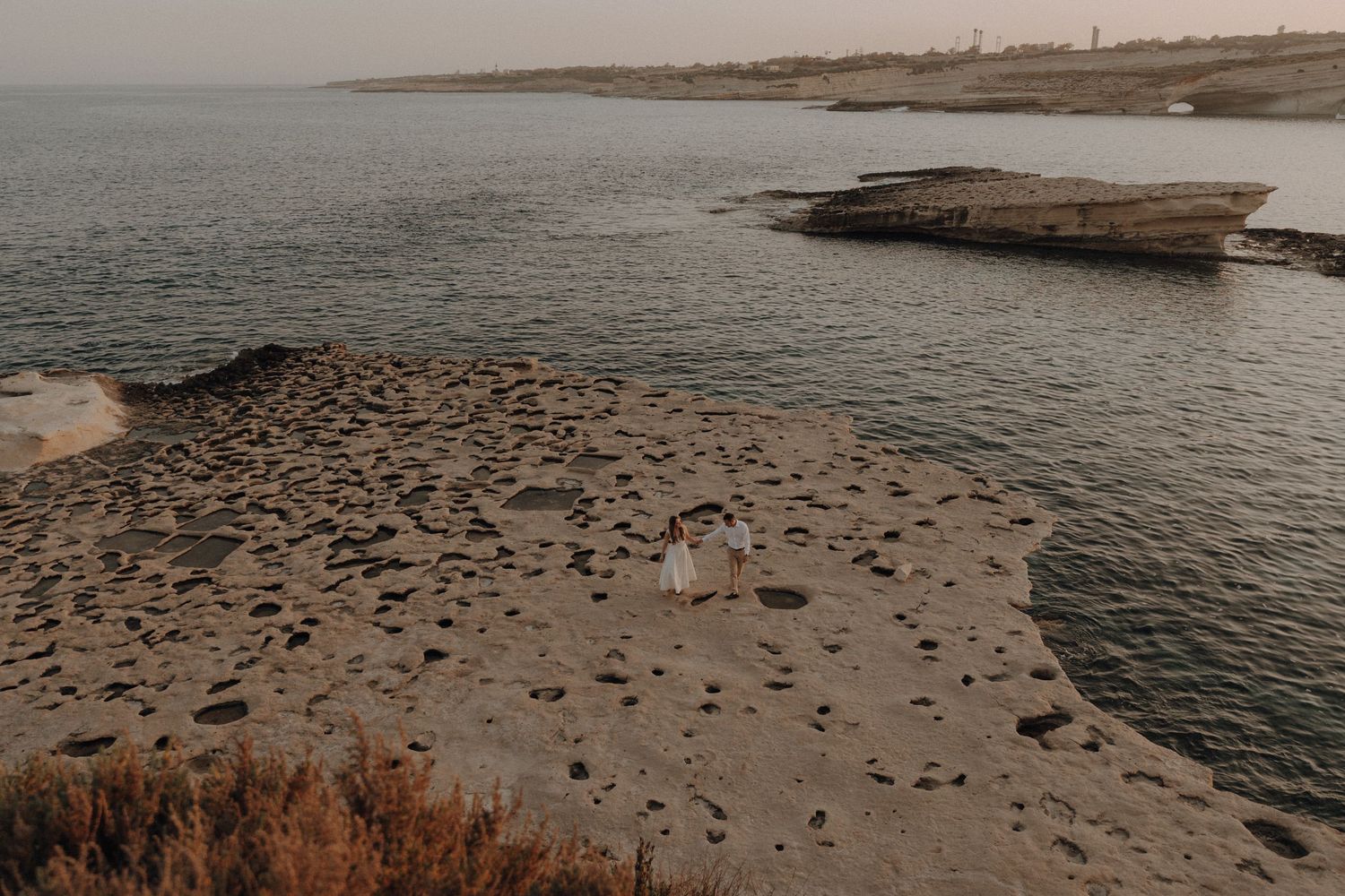 Aerial view of a couple standing on rocky coastal shoreline at dusk with calm ocean waters surrounding them.