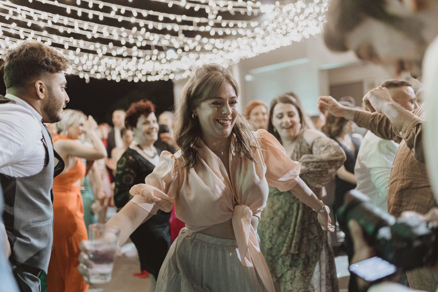 Party guests dance under decorative string lights at an indoor reception.