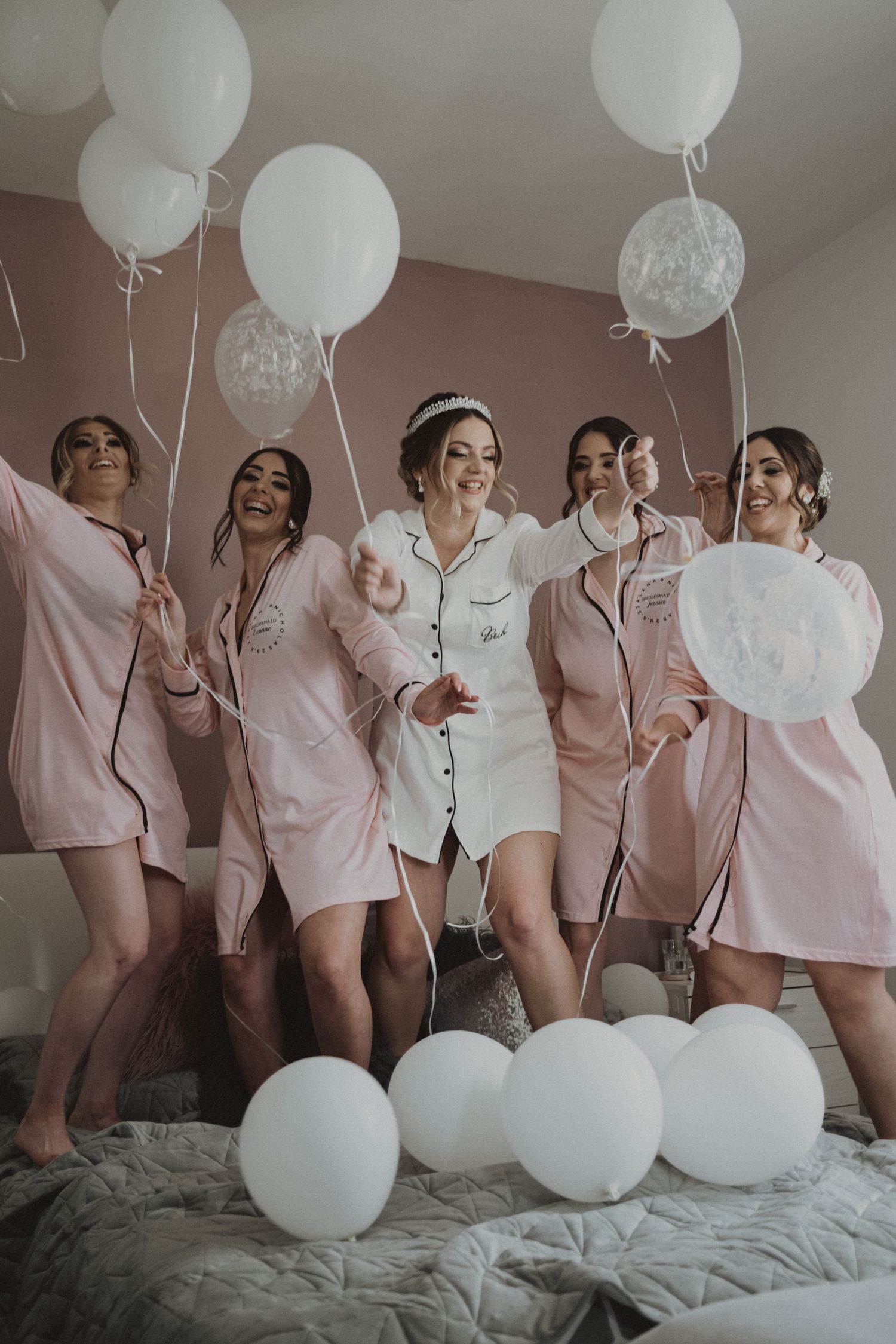 Wedding party in matching pink pajamas having fun with white balloons on a bed during pre-ceremony preparations.