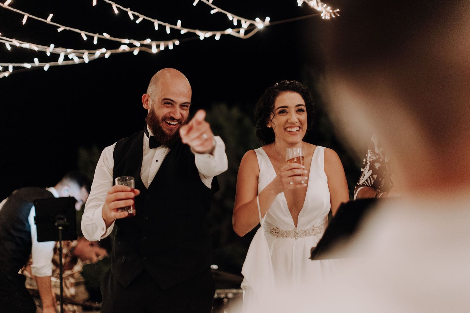 A couple raises their glasses in a wedding toast under twinkling string lights.