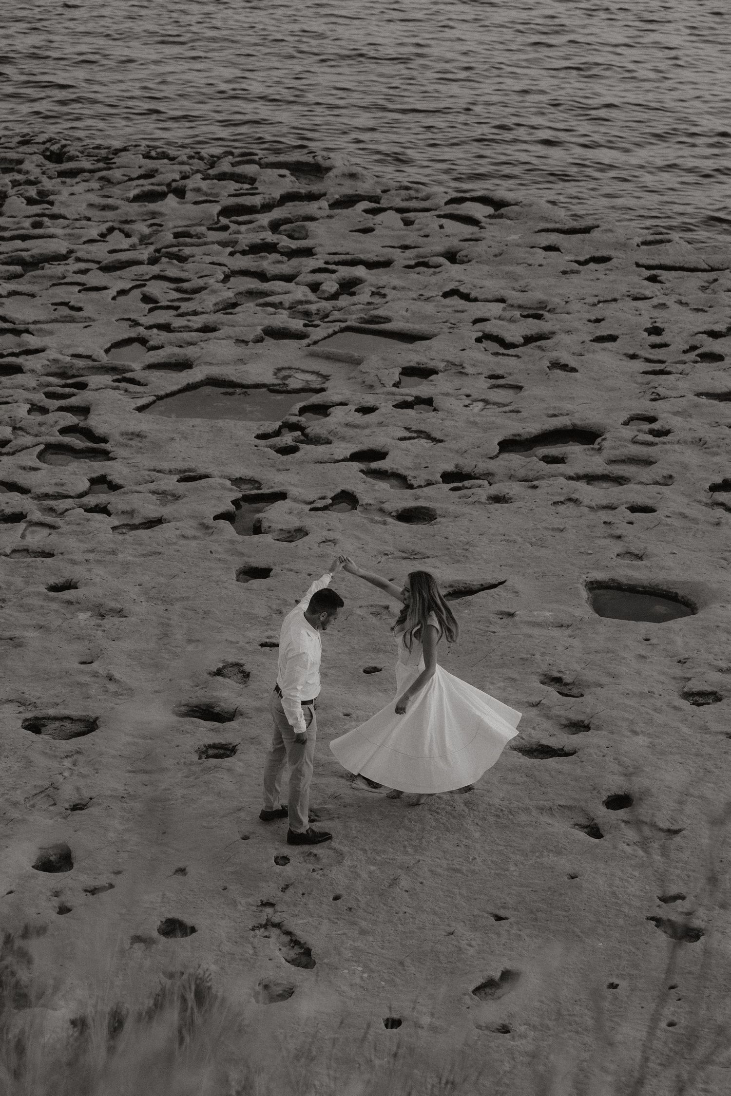 Black and white aerial photo of a couple dancing on a rocky beach, woman in flowing white dress.