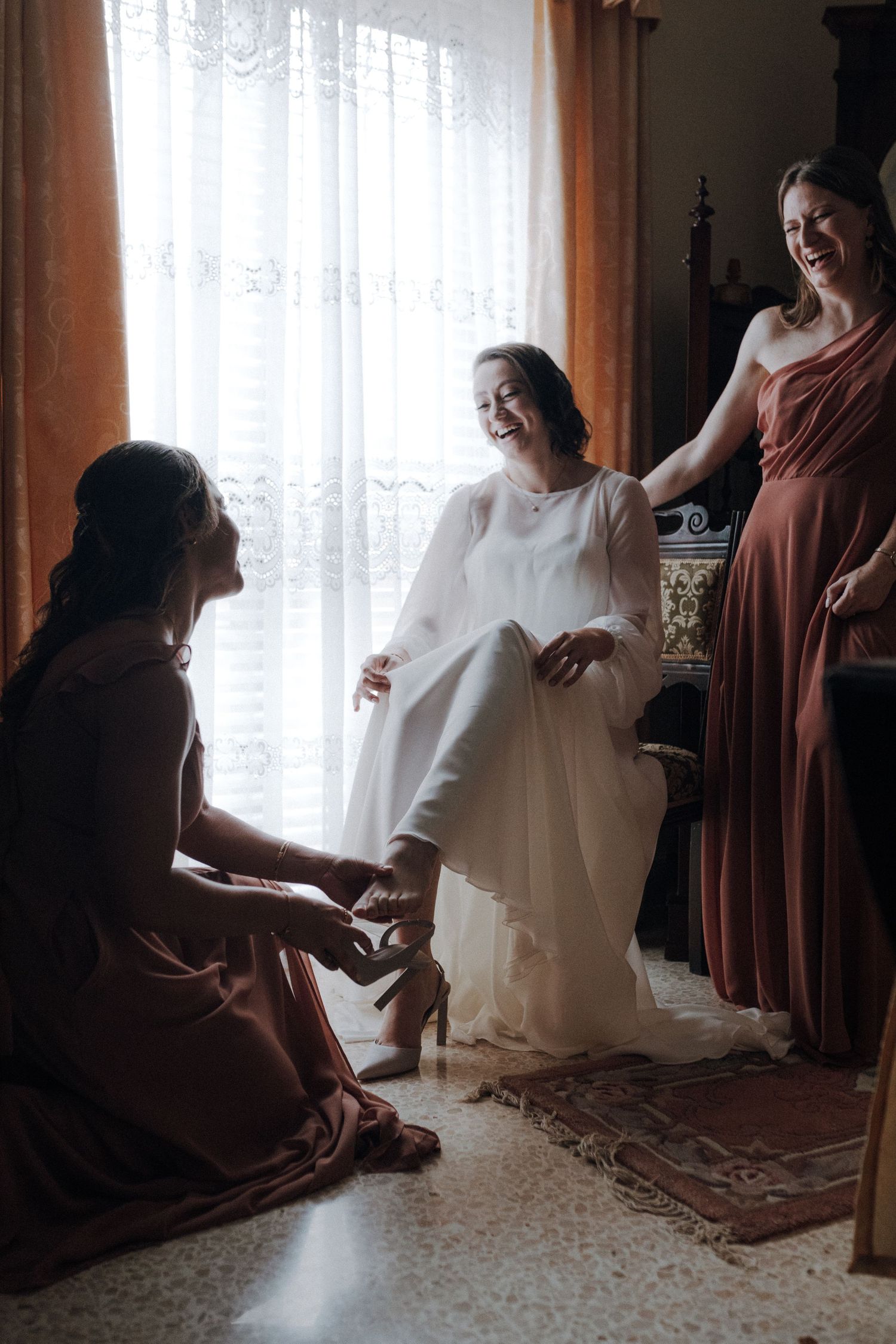 Three women share a candid moment during wedding preparations in a sunlit room.