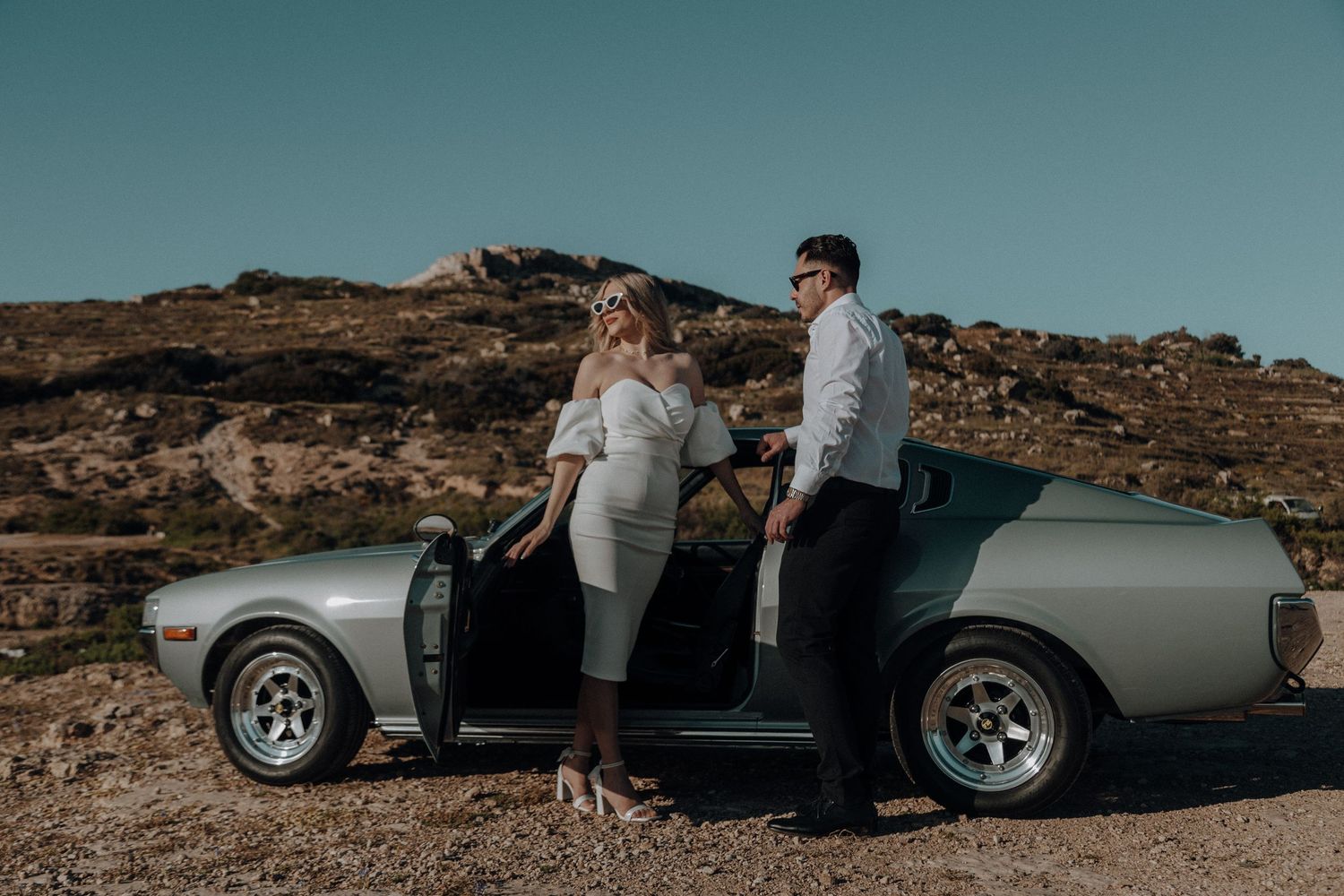 A classic silver sports car poses against a scenic desert landscape backdrop during a photo shoot.