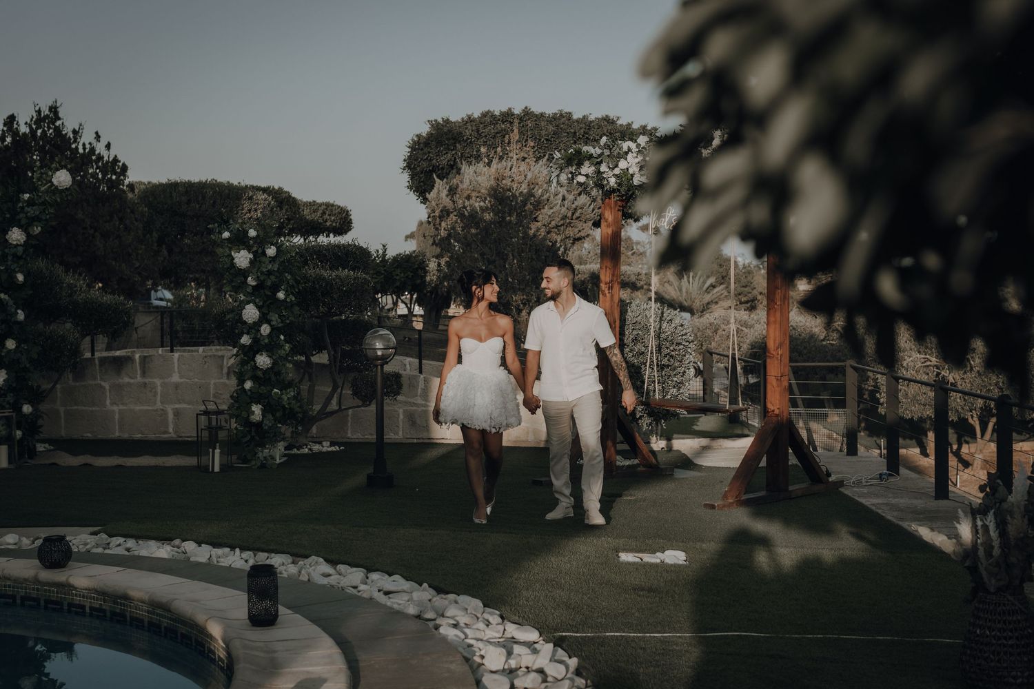 A couple in white outfits walking together along a poolside path at dusk.