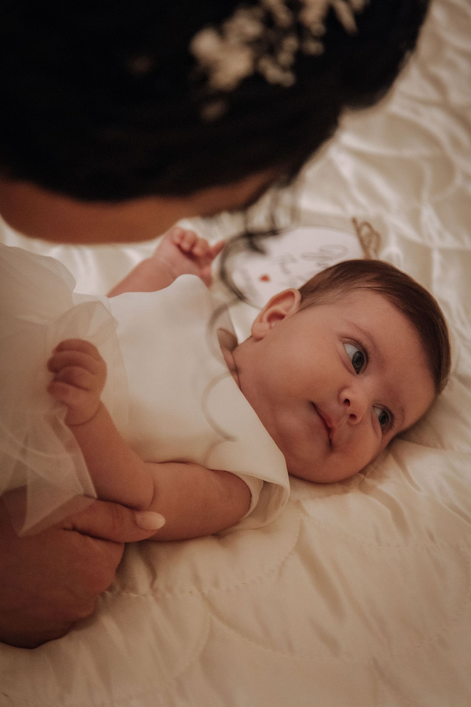 A baby in white clothing lies on white bedding looking up with bright eyes.