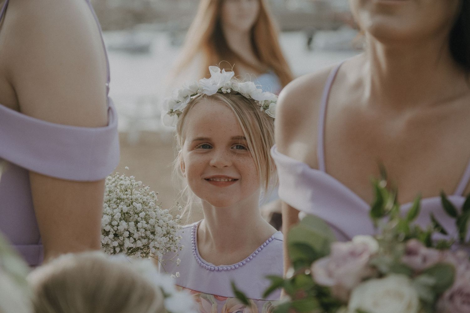 Young attendant in floral crown and lavender dress smiles during outdoor wedding ceremony.