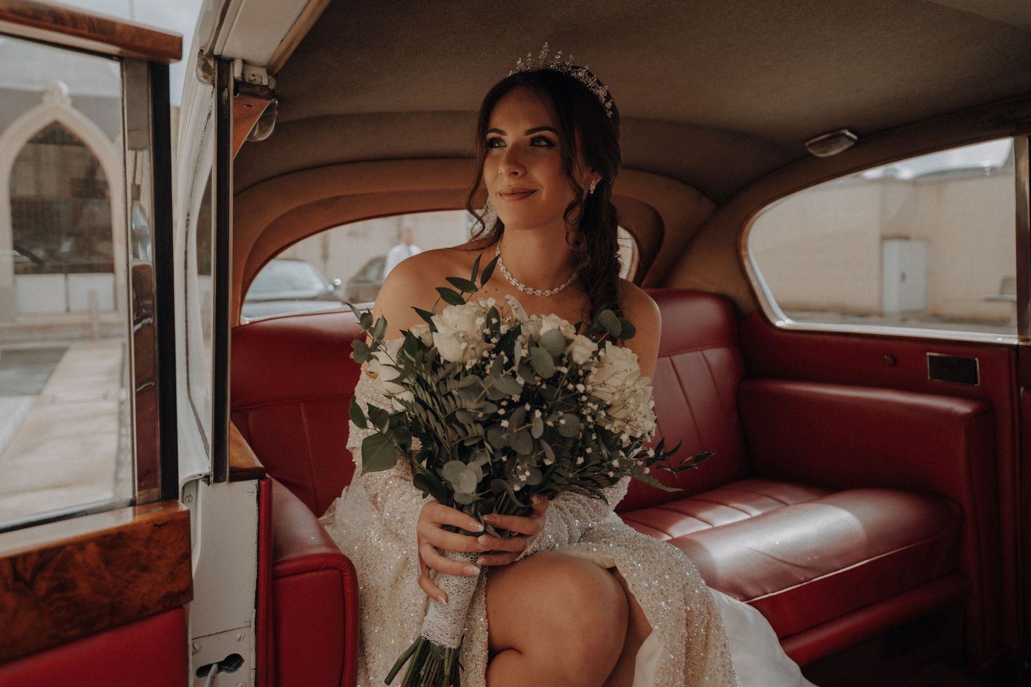 Bride in vintage car holding bouquet wearing crystal crown and off-shoulder dress.