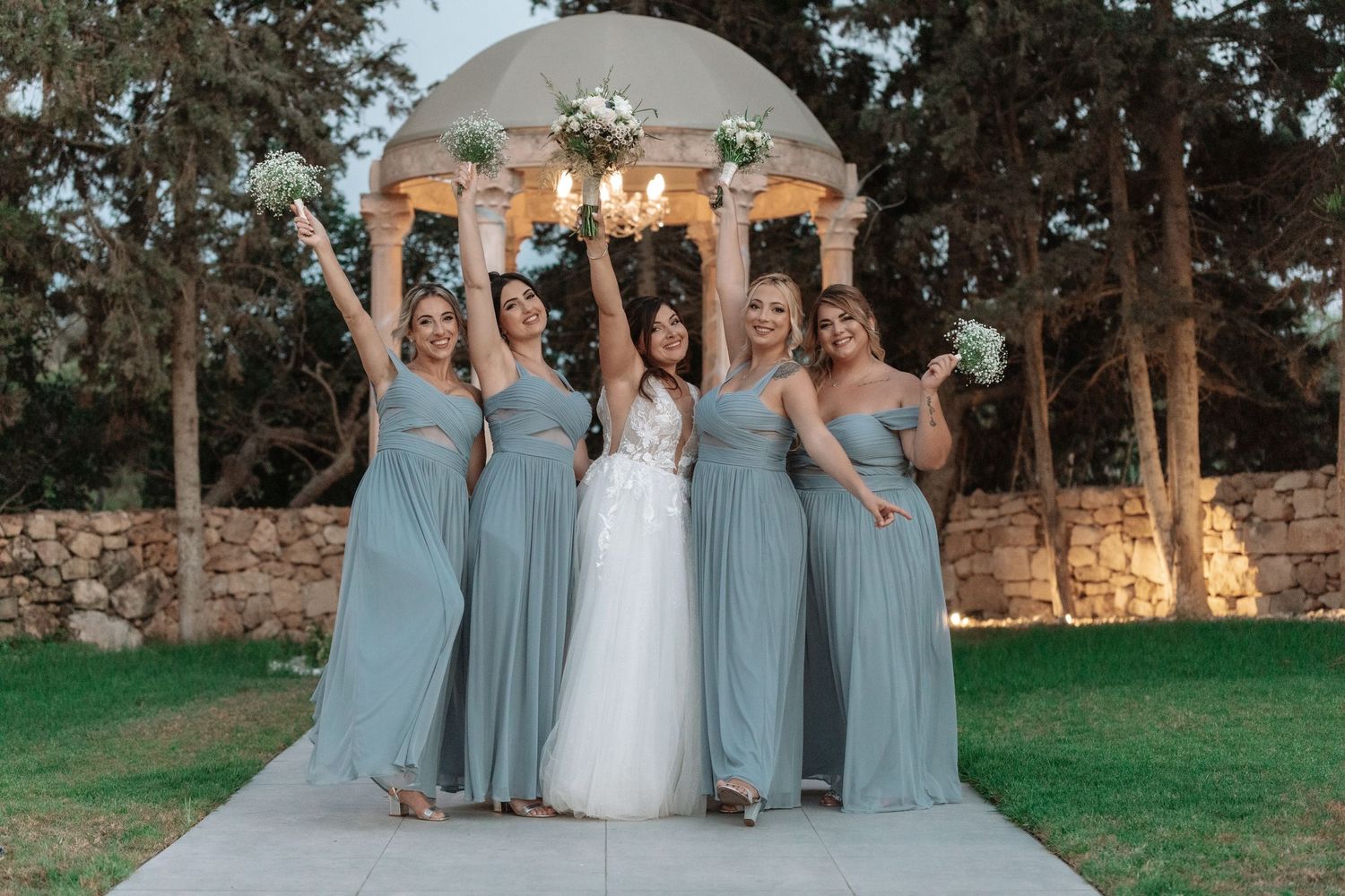 Bridesmaids in light blue dresses celebrate joyfully with the bride in front of an ornate gazebo.