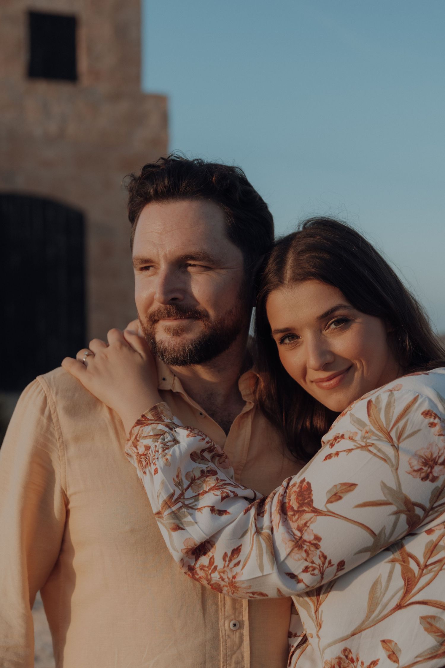 A romantic couple embraces during golden hour on a beach, with the woman wearing a floral print dress.
