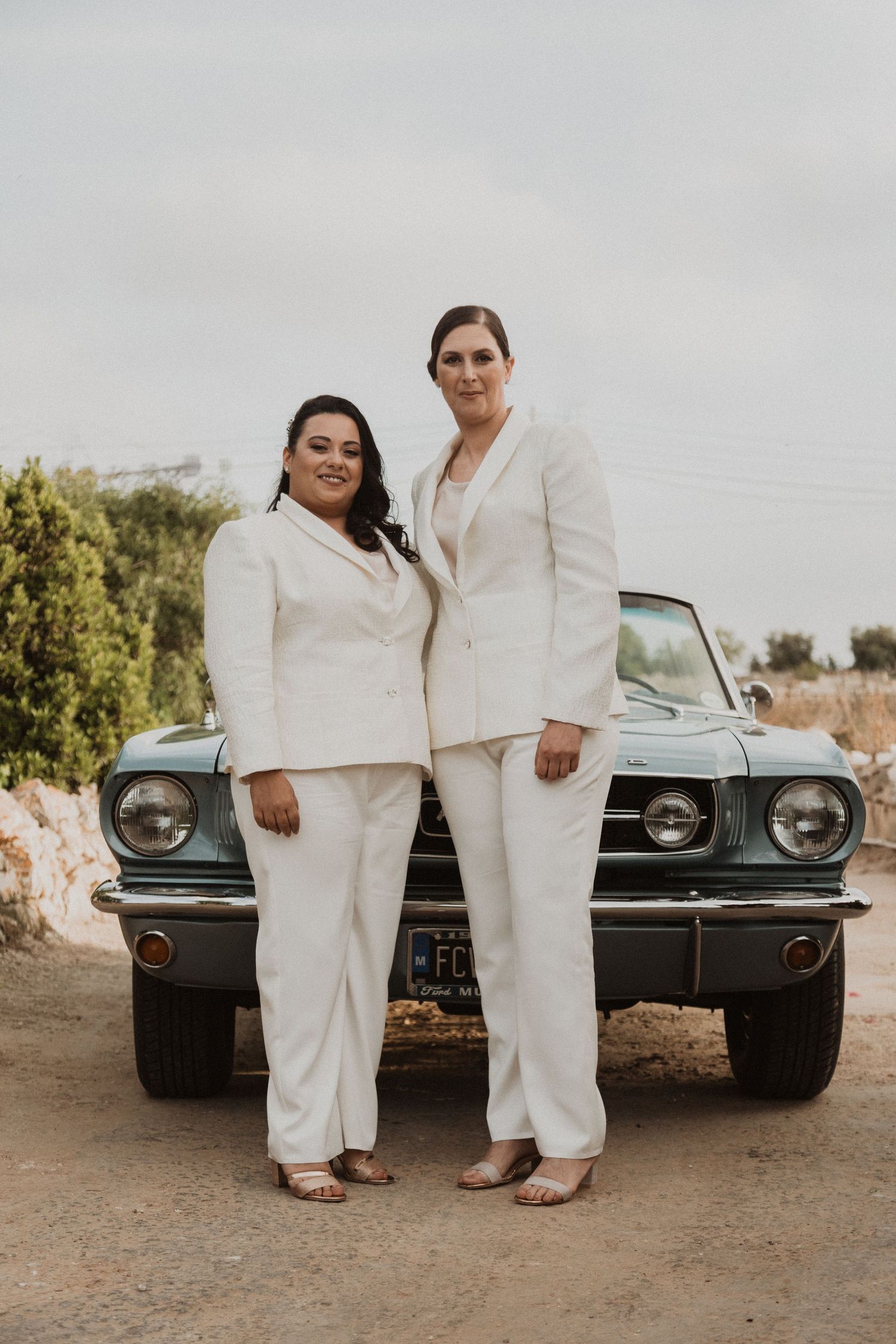Two people in white outfits posing in front of a classic car.