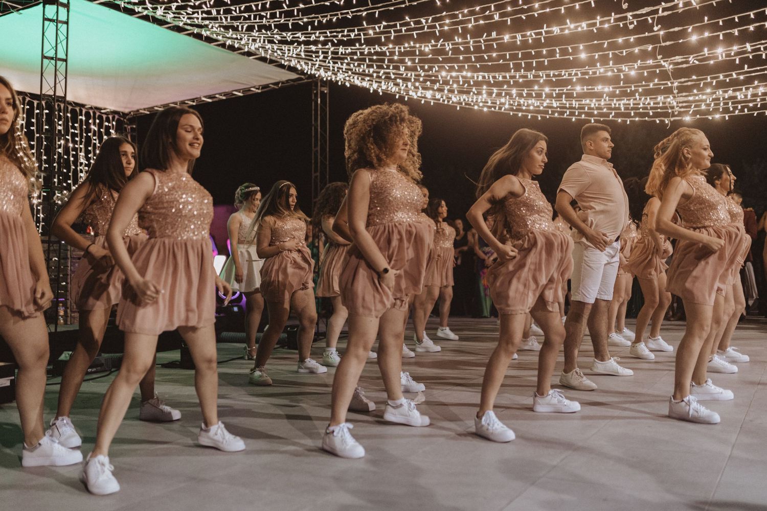 Group performing a synchronized dance in matching blush pink dresses and white sneakers under string lights.
