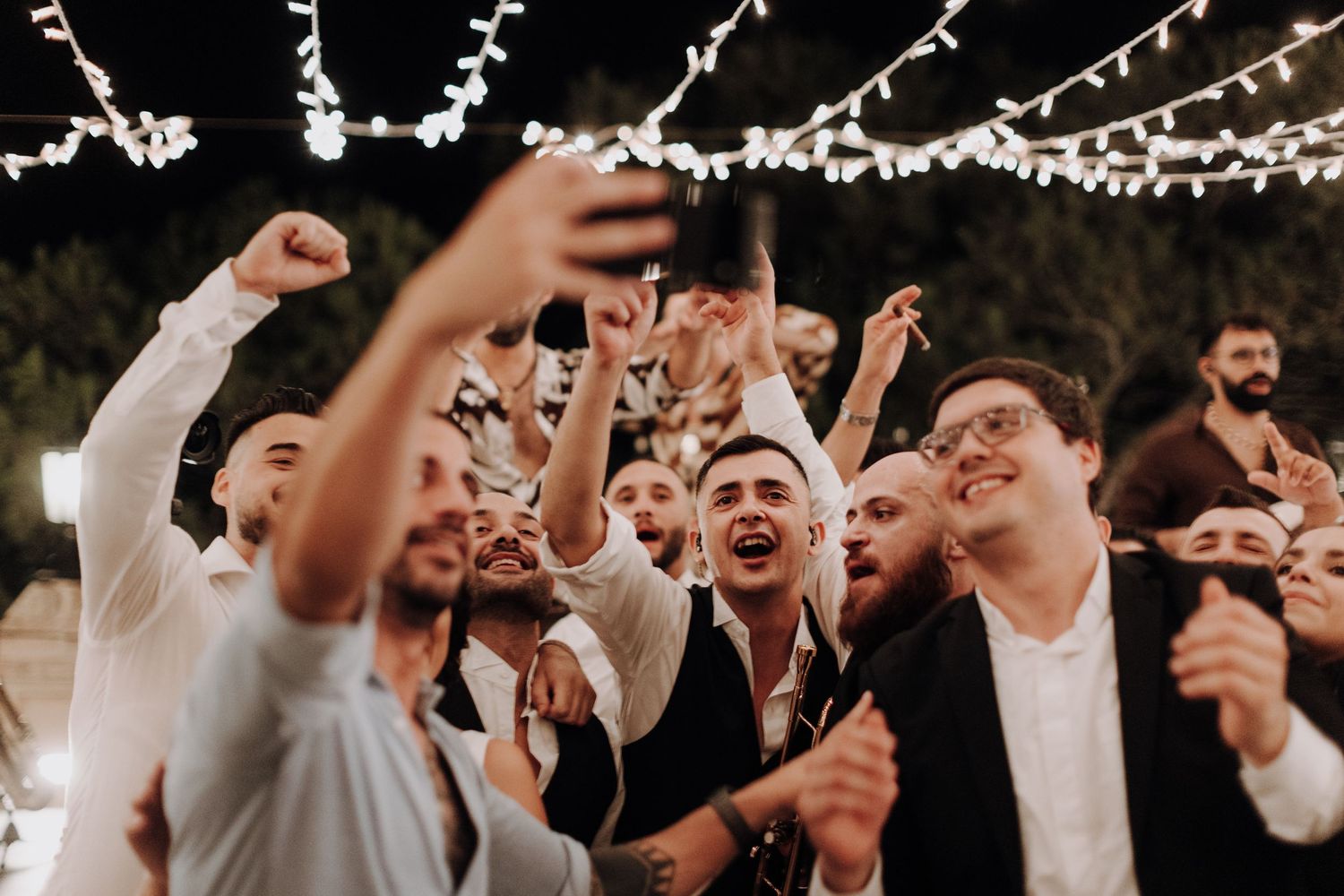 Energetic wedding guests dance and celebrate under string lights at an outdoor reception.