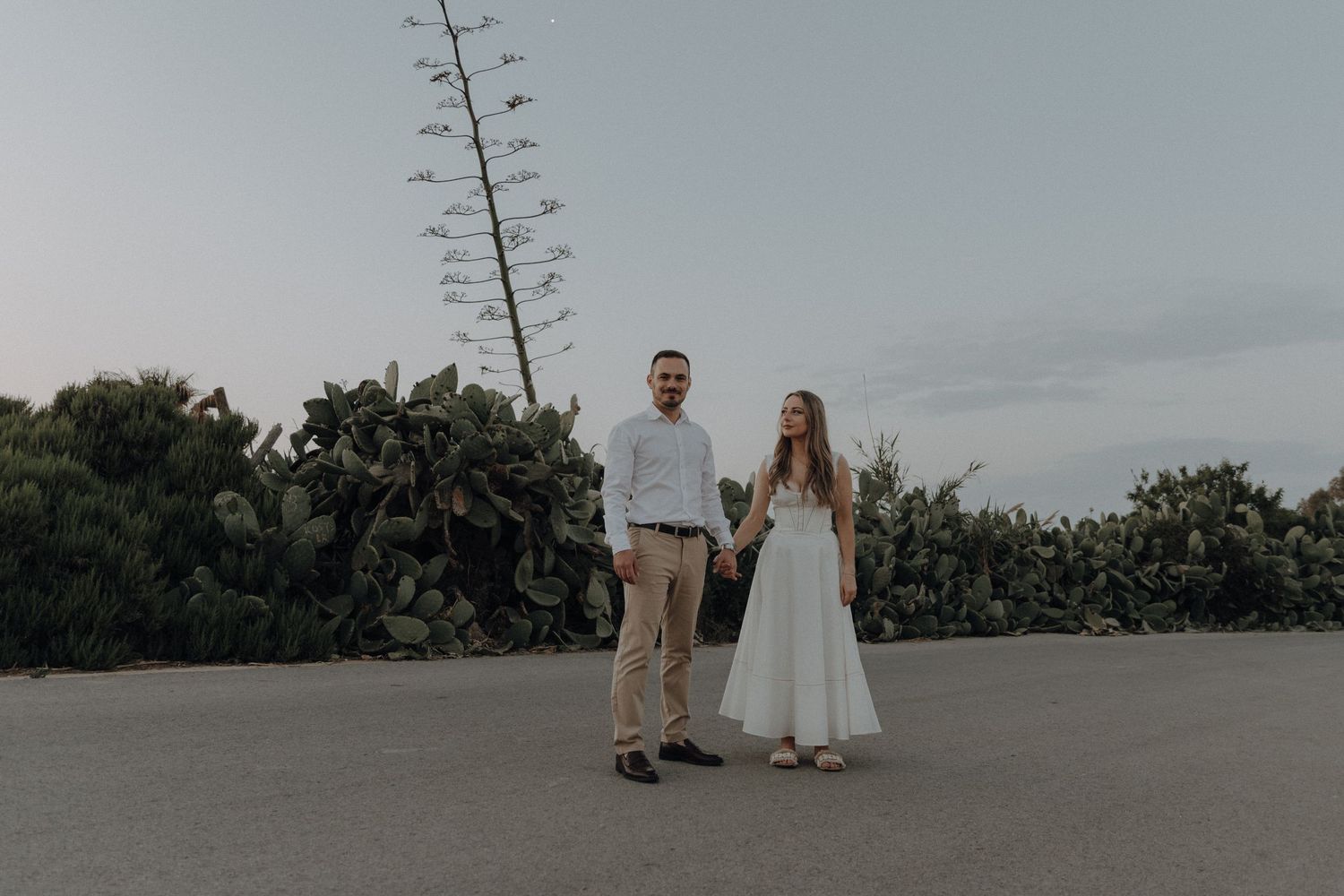 Couple holding hands outdoors at dusk with cacti and tall agave plant behind them.