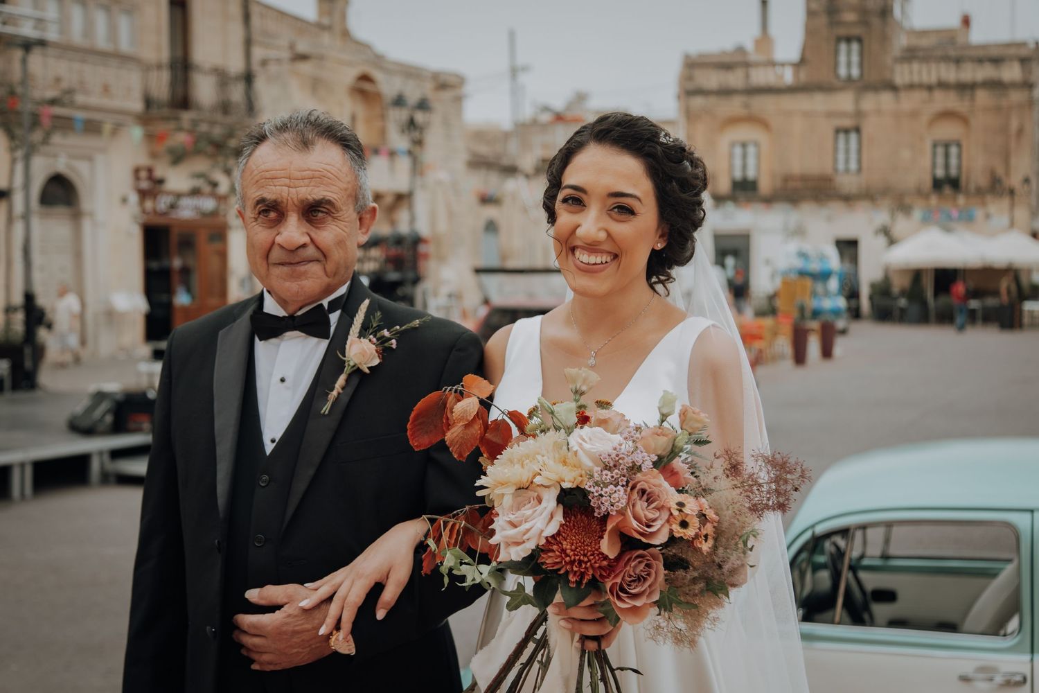 Bride holding floral bouquet stands with companion outside historic stone architecture.