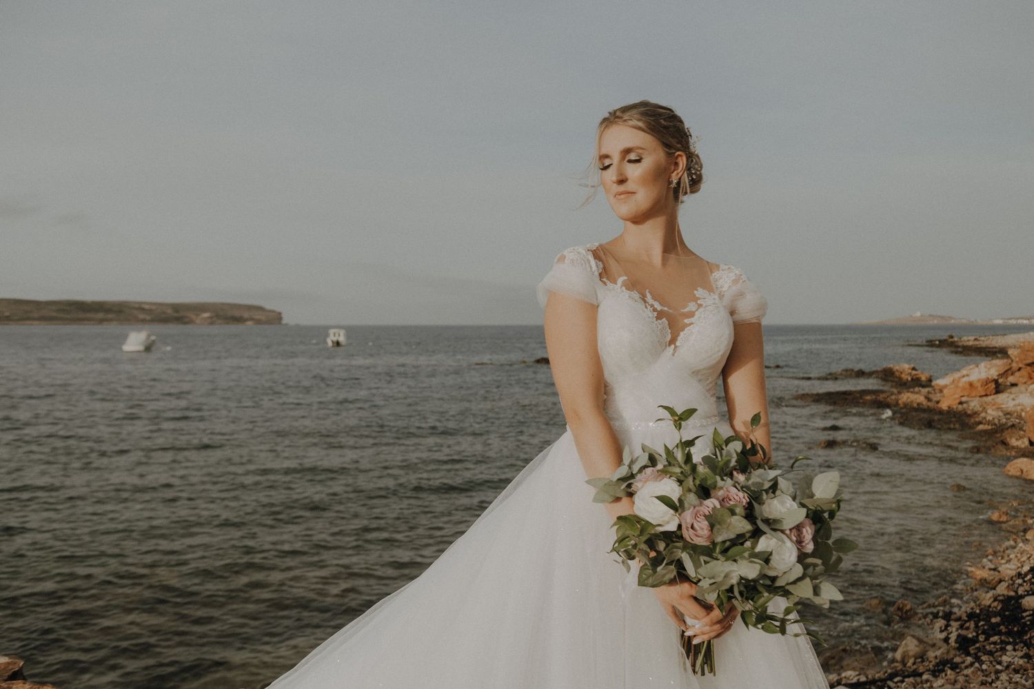 Elegant bride holding romantic bouquet poses on rocky shore during golden hour.