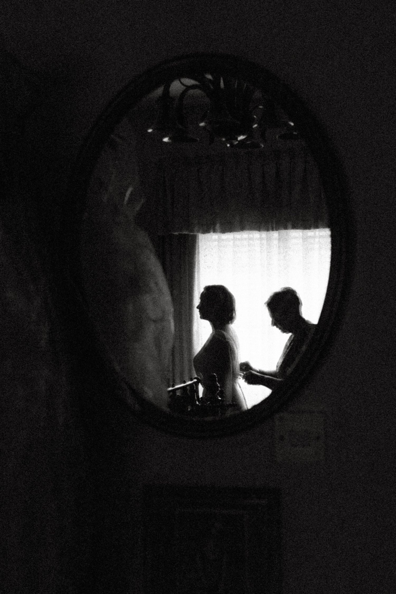 Artistic black and white reflection in oval mirror showing silhouettes near a window.