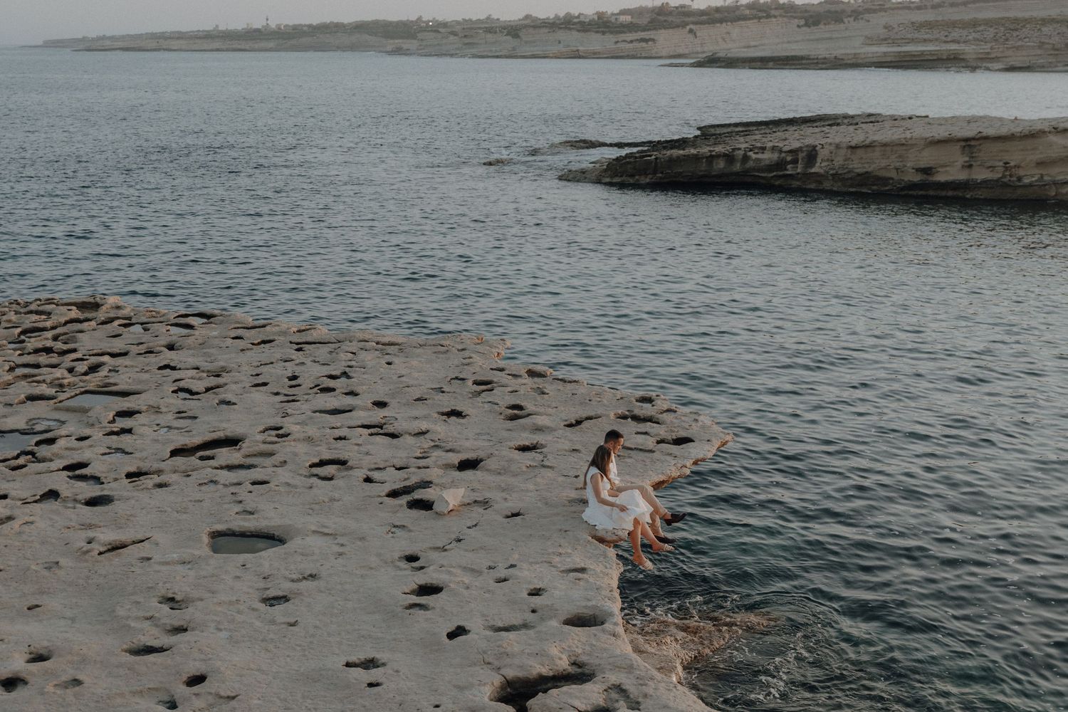 Woman in white dress sitting on rocky shoreline edge overlooking calm ocean waters.