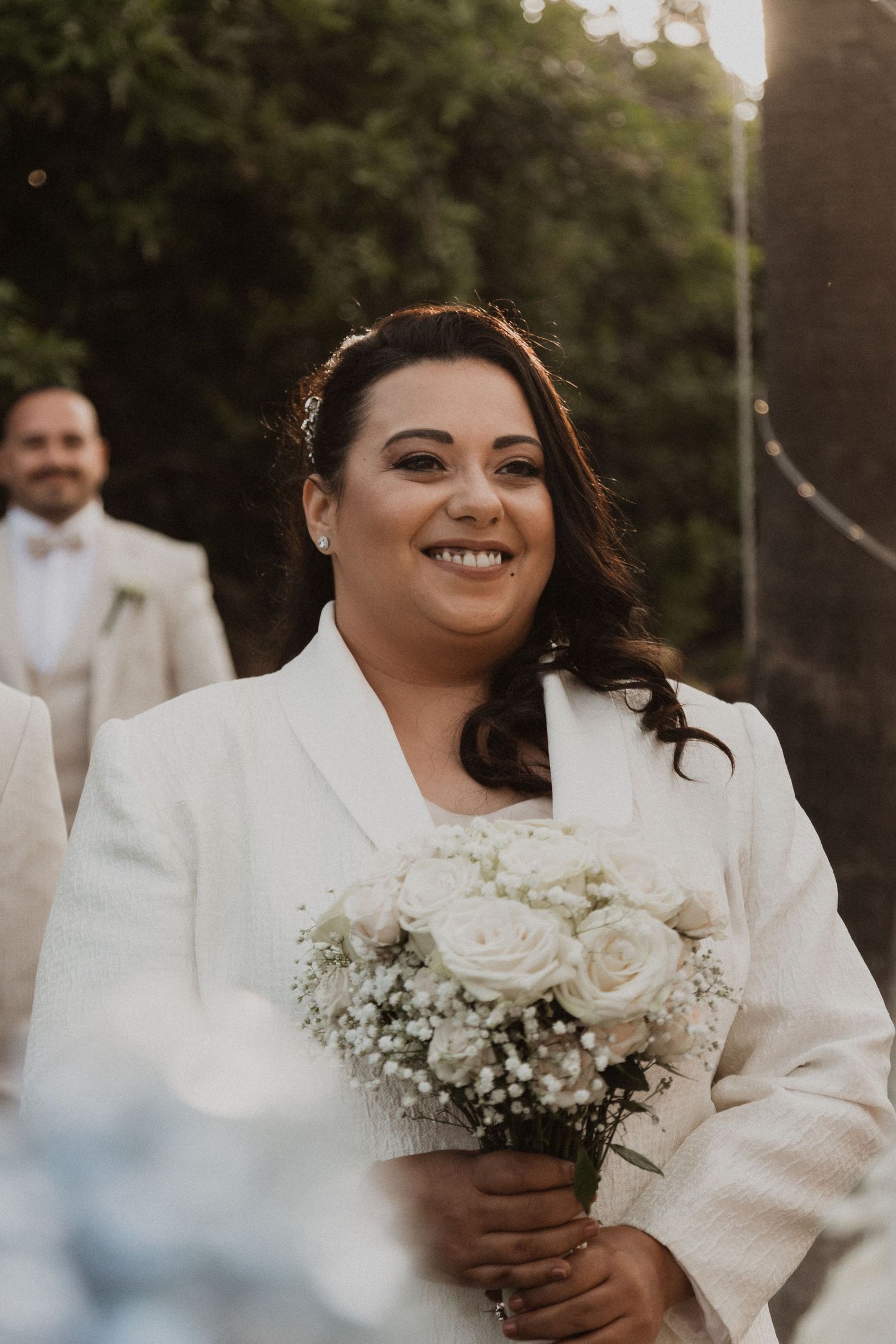 A joyful bride in a white wedding suit holds a bouquet of white roses and baby's breath on her special day.