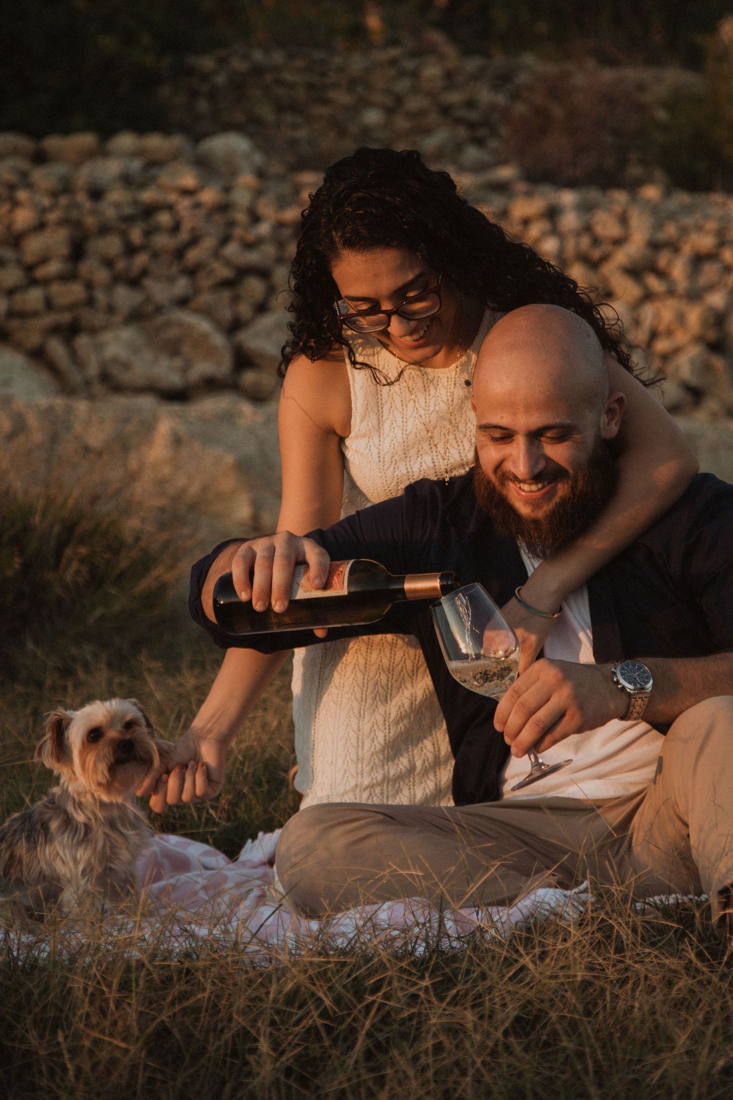 A couple sharing wine during a sunset picnic against a rustic stone wall backdrop.