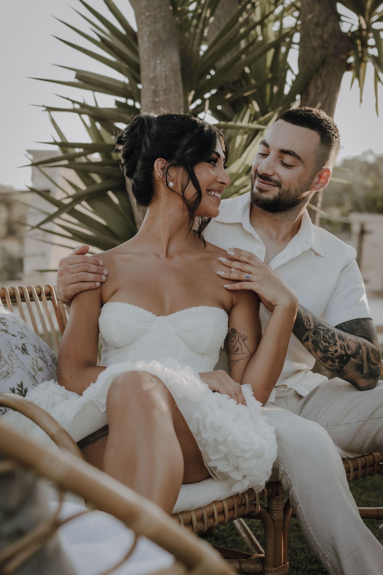 A romantic couple sharing an intimate moment on a wicker chair surrounded by tropical plants.