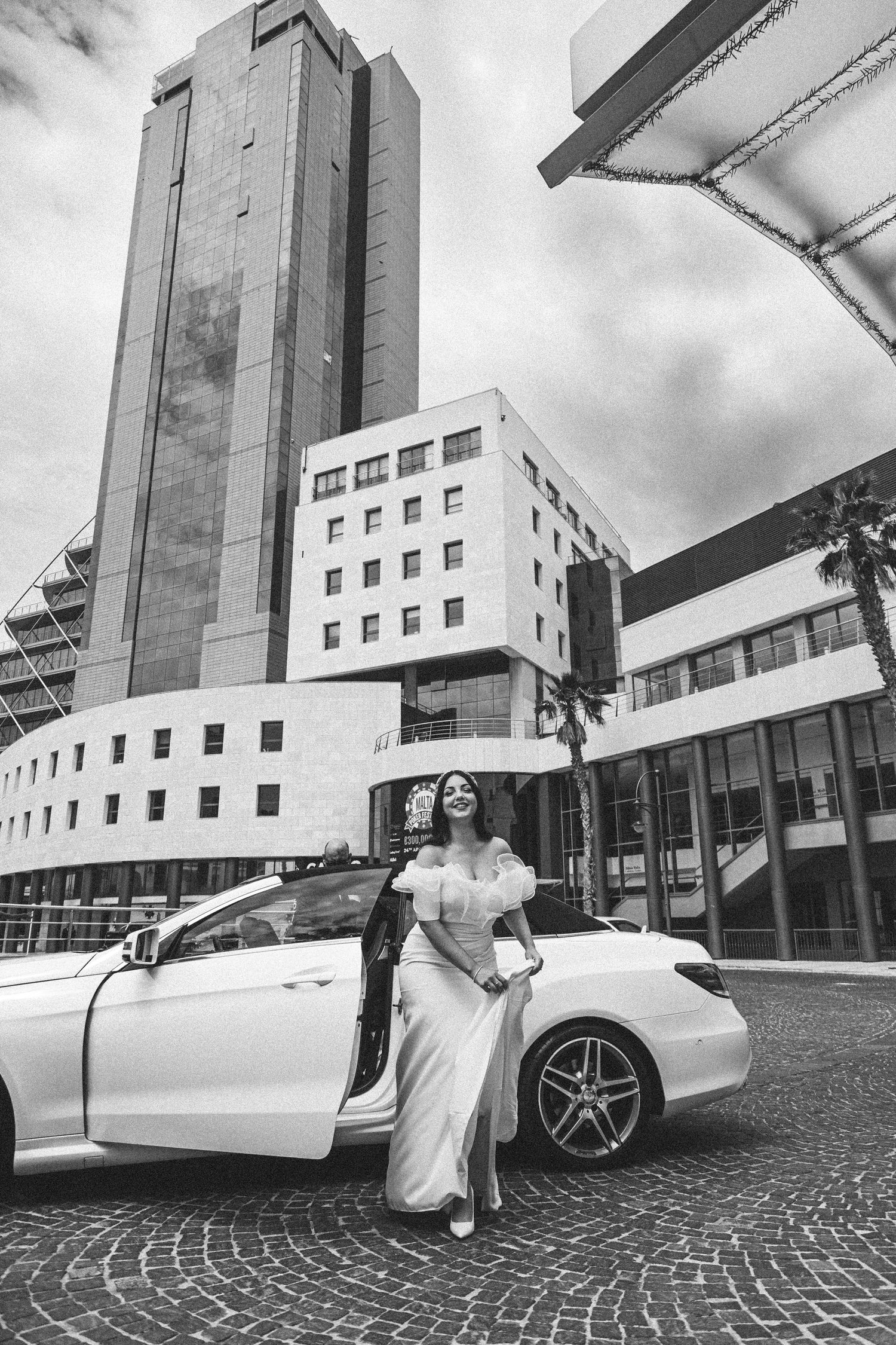 A bride in a white gown poses elegantly next to a luxury car outside a modern building.