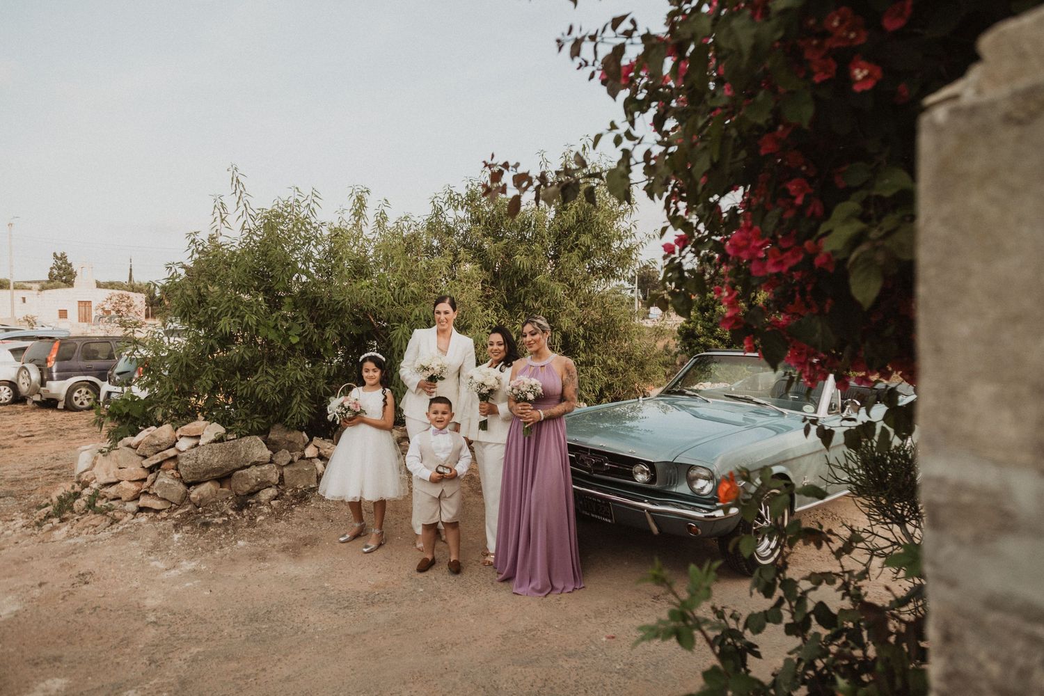 Wedding party gathered by vintage car against stone wall with flowering vines.
