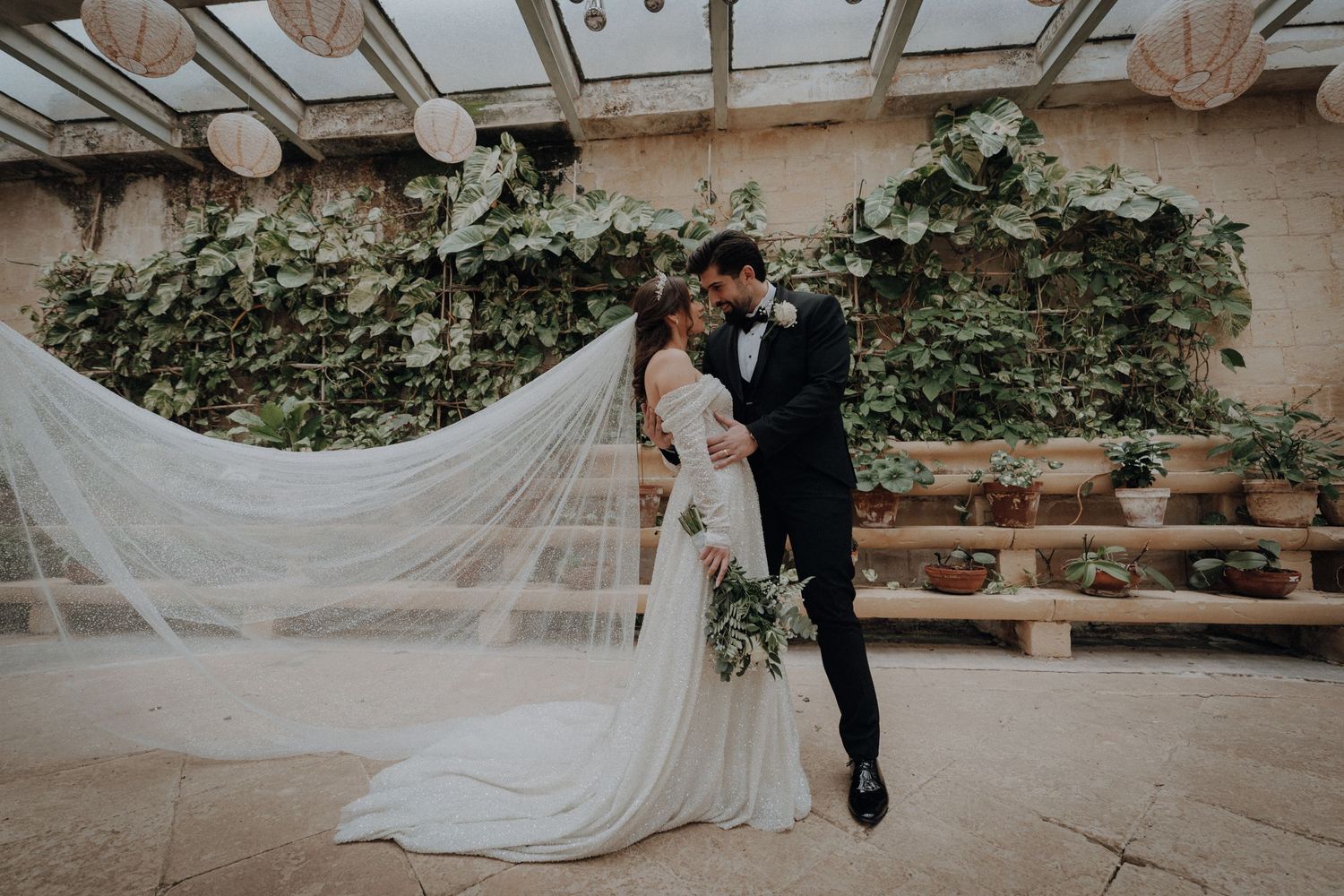 A long white wedding veil flows dramatically in the wind against a rustic stone backdrop at sunset.
