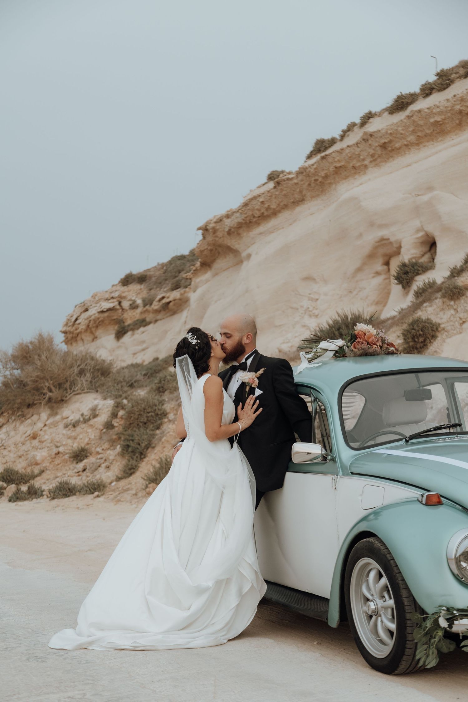 Newlyweds embrace against vintage blue Volkswagen Beetle on coastal cliff.