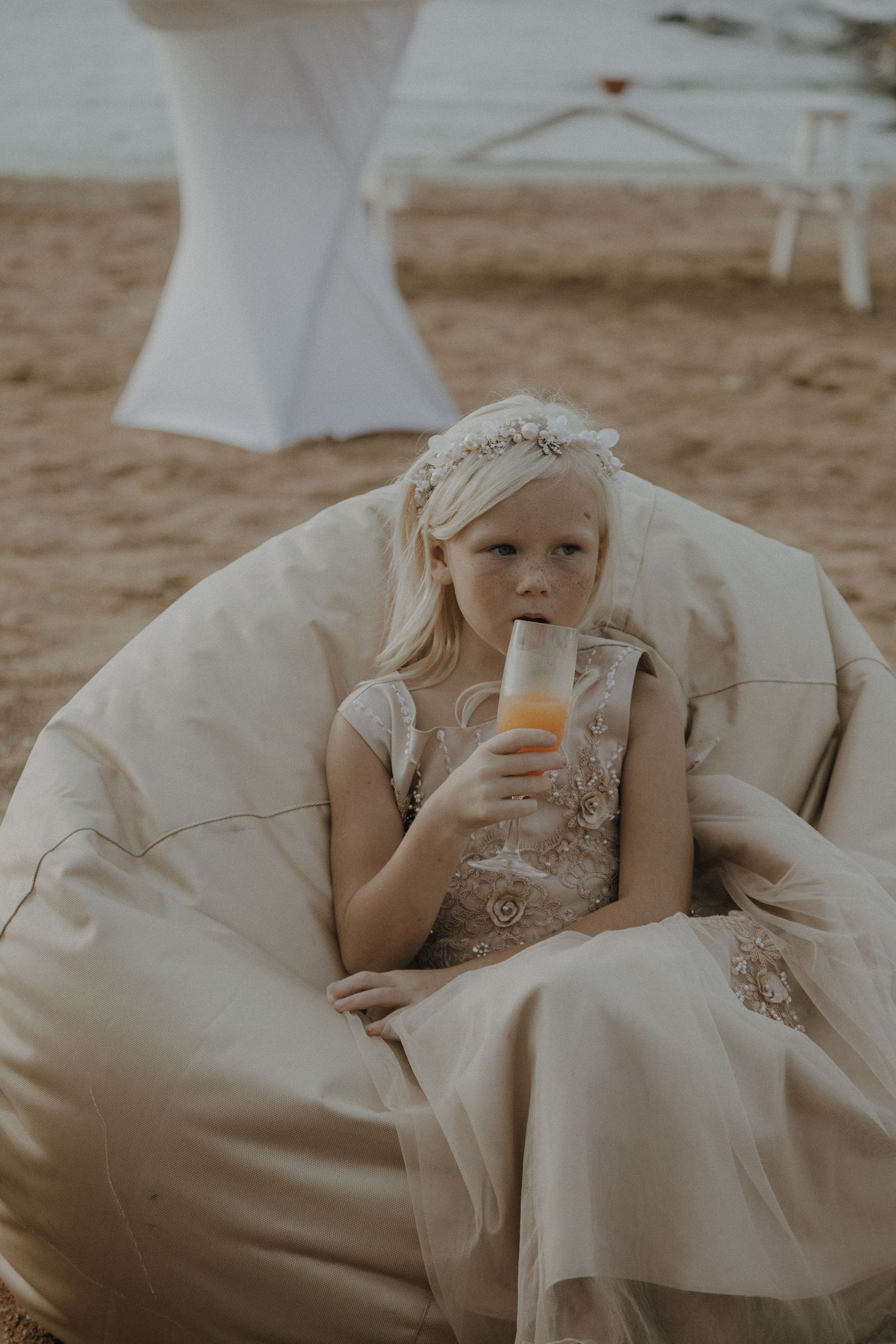 Young wedding guest in formal dress relaxes with drink in beanbag chair on beach.