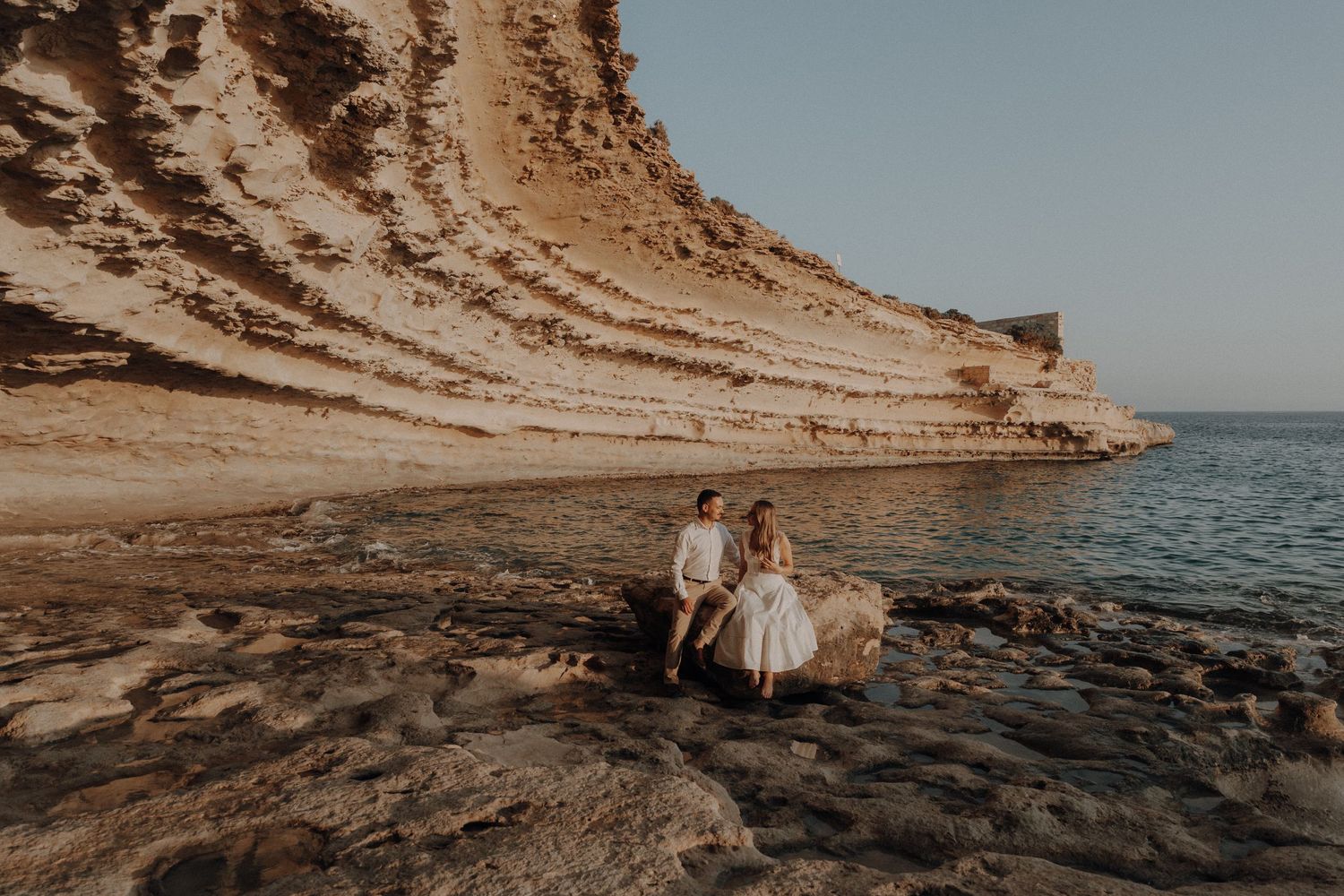 Couple sitting on rocks by a stunning coastal cliff at golden hour during a romantic beach photo session.