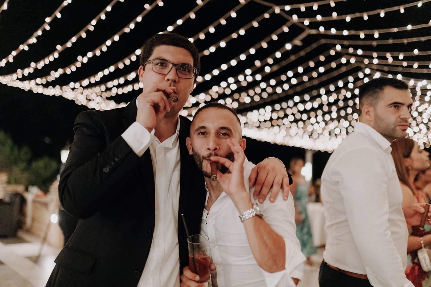 Wedding guests share a playful moment under decorative string lights during the reception.