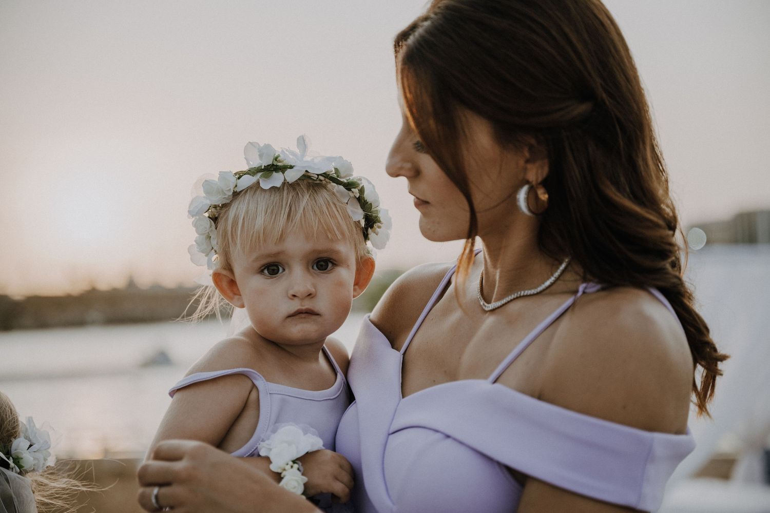 Mother in lavender dress holds toddler wearing floral crown by waterfront during sunset.
