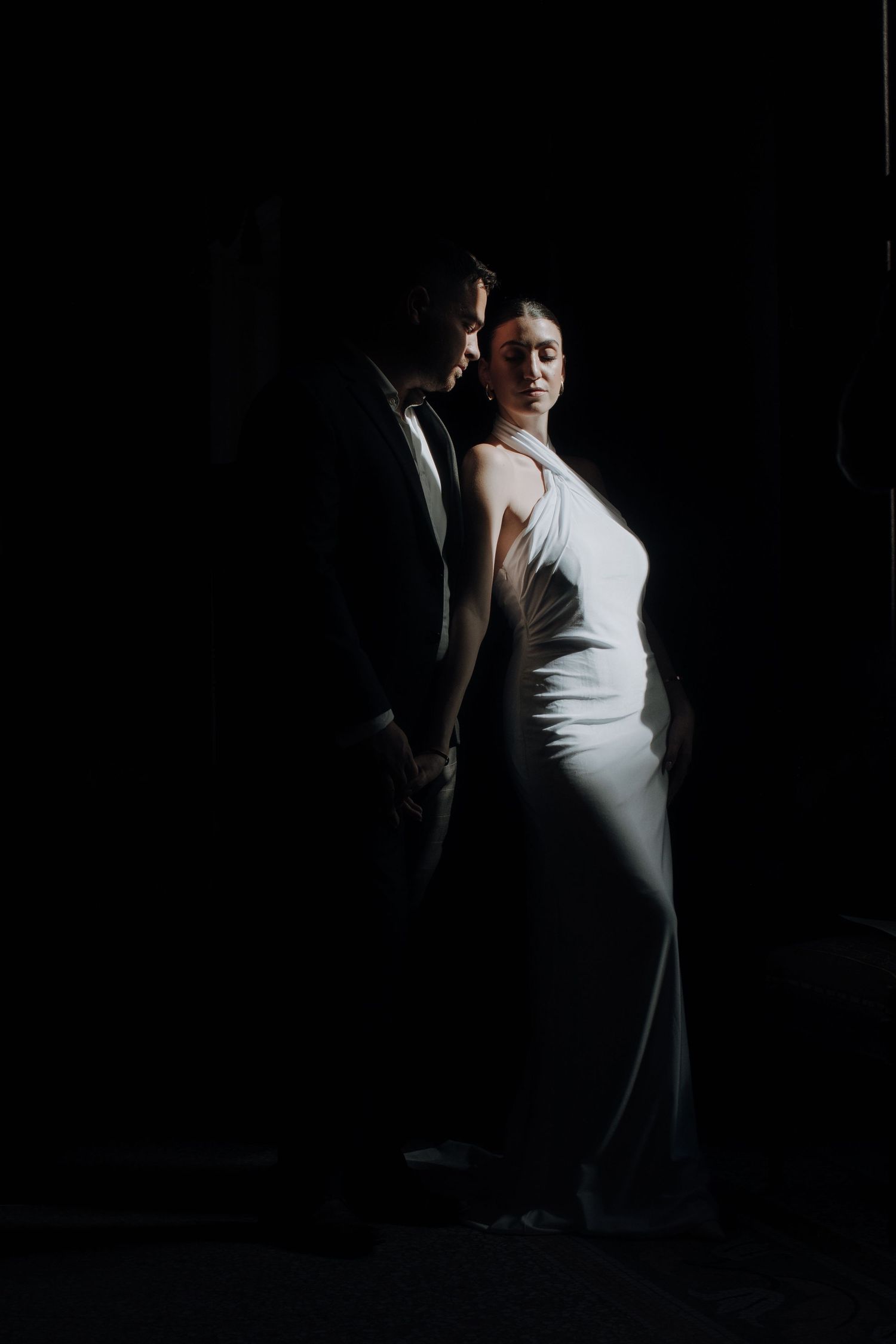 Dramatic portrait of couple in formal wear with strong contrast lighting against dark background.