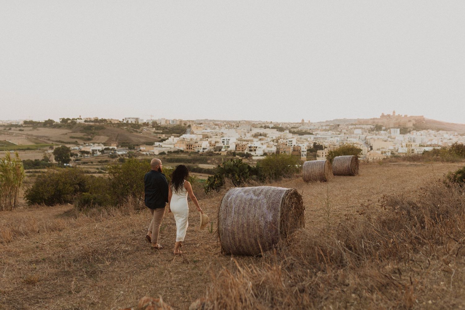 Two people stroll through a rustic field with hay bales and a Mediterranean town in the distance.
