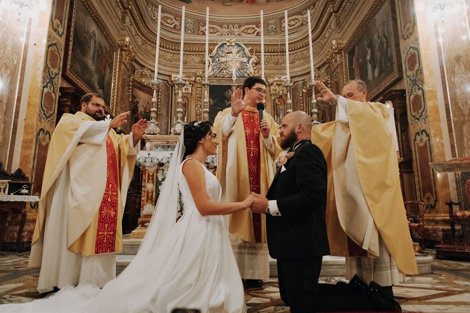 A wedding ceremony takes place at an ornate church altar surrounded by clergy in white robes.