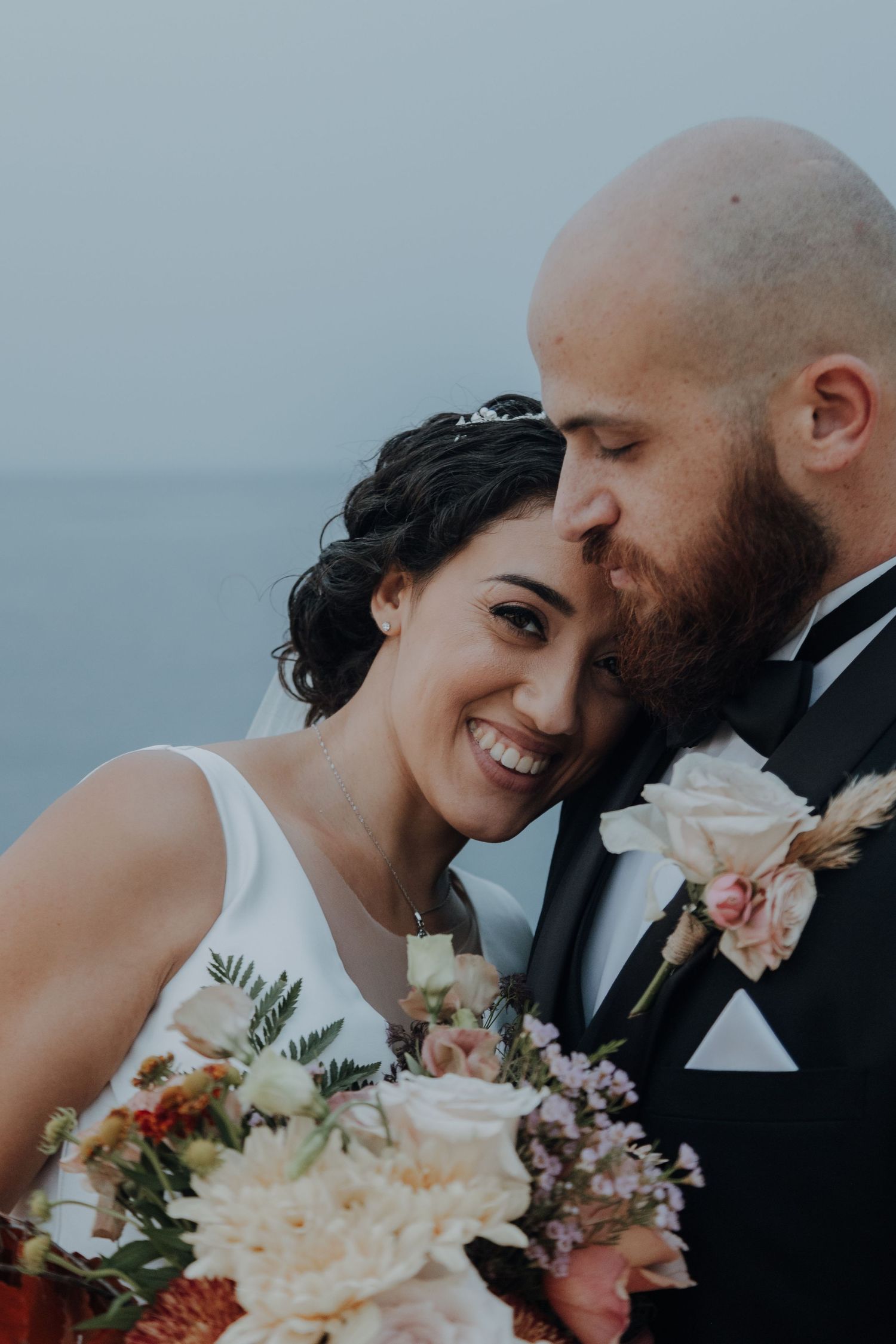 A romantic wedding portrait showing an intimate moment between newlyweds with a coastal backdrop.
