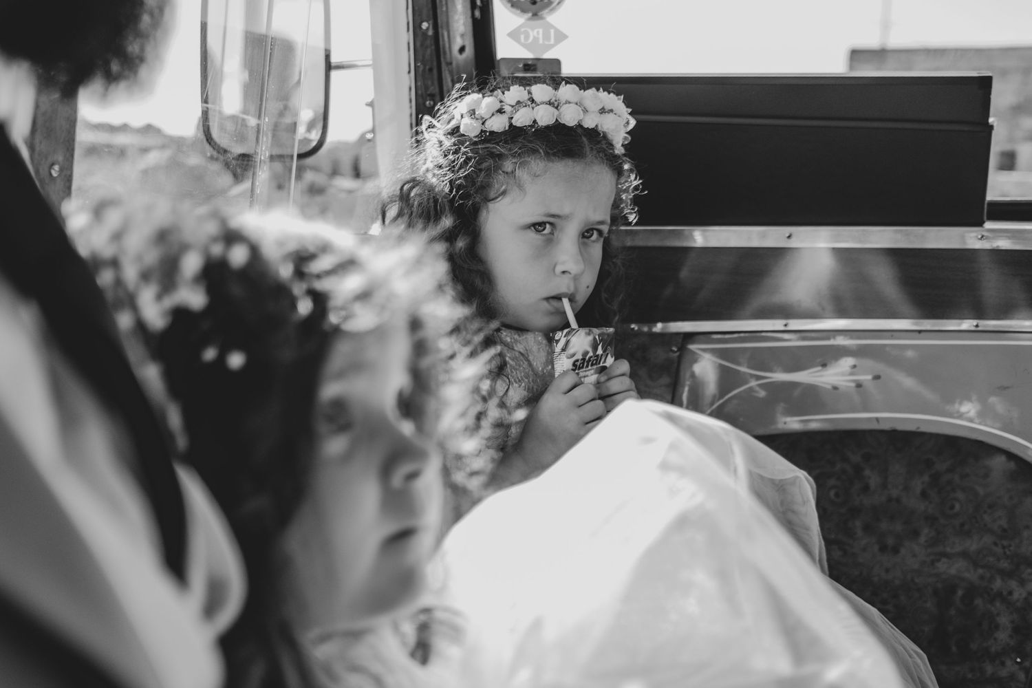 Young flower girl in white dress with floral crown looks pensive on bus seat.