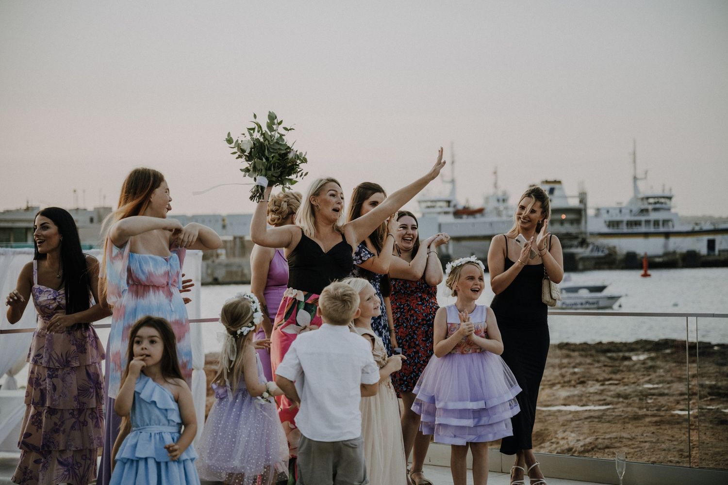 A group of wedding guests celebrating during the bouquet toss by the harbor.
