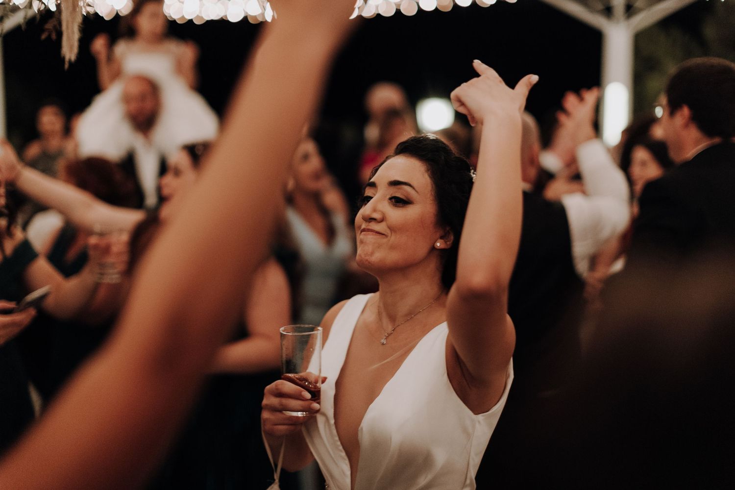A bride dances with raised arms under string lights during her wedding reception celebration.